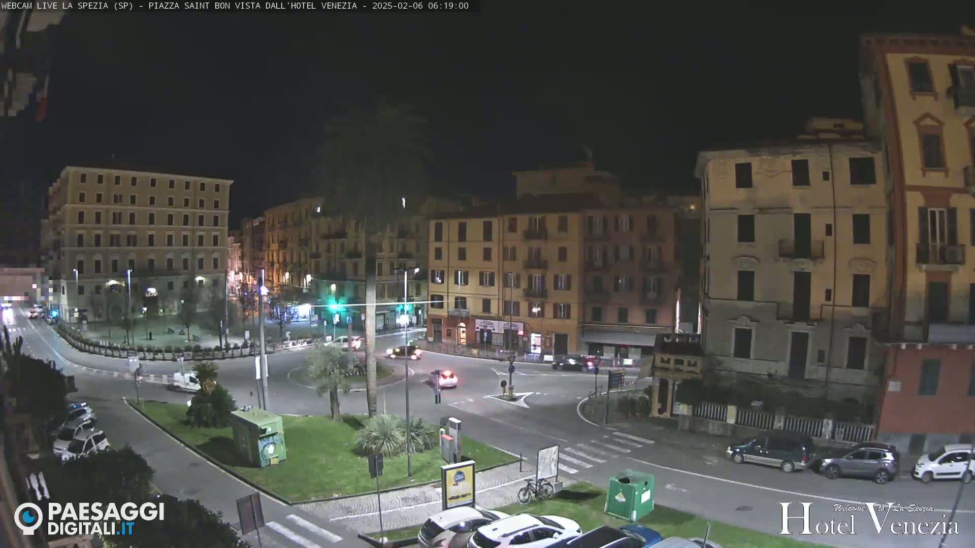 A nighttime view of a city square with several multi-story buildings surrounding a roundabout with cars and a few pedestrians.