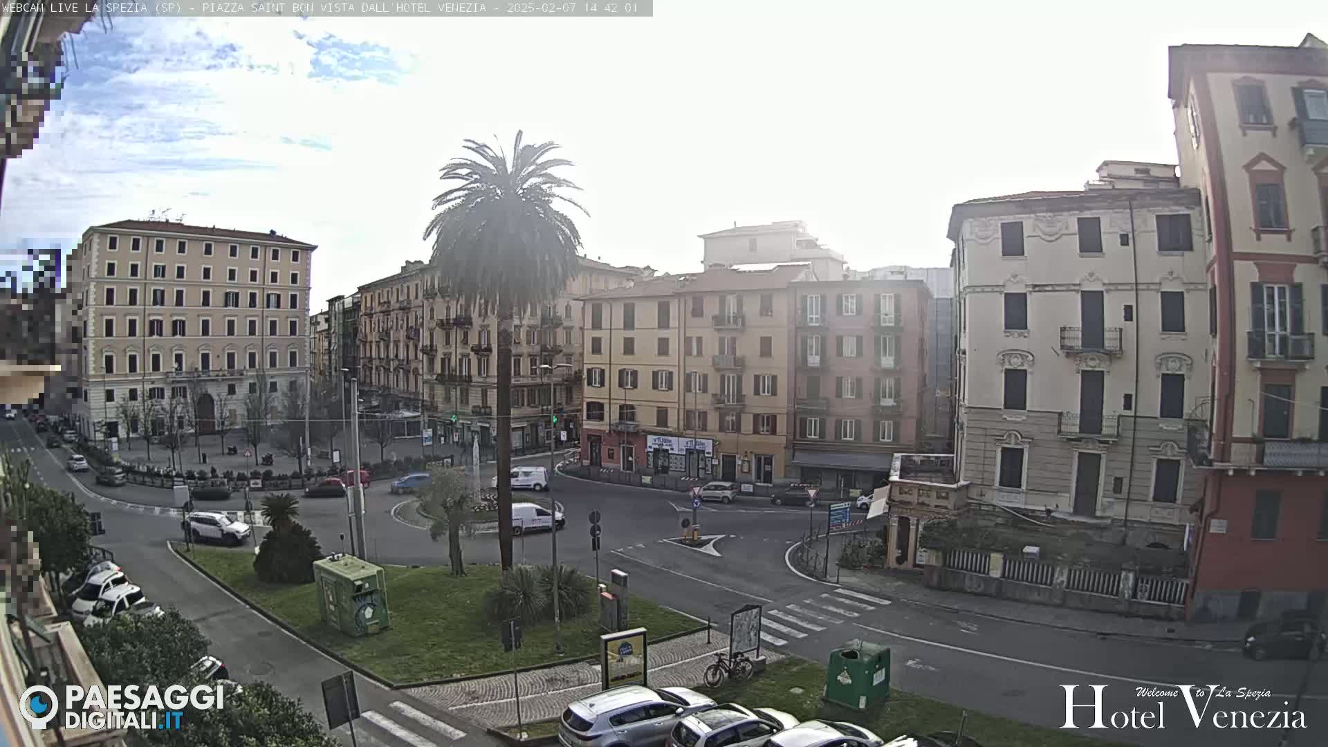 A mostly sunny day in a European city square, with several multi-story buildings surrounding a roundabout where cars are parked and driving.