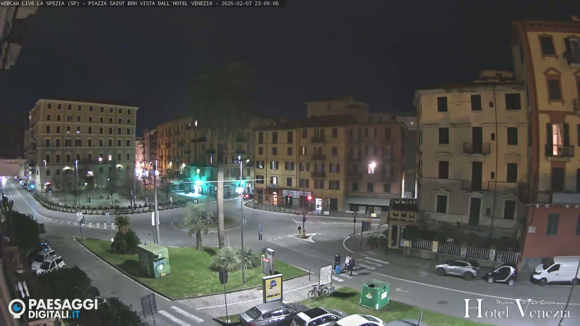 A nighttime view of a town square in a European city, featuring several multi-story buildings, a small roundabout, and sparse traffic.