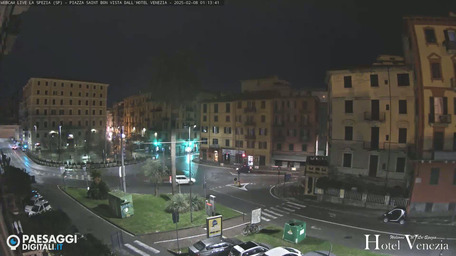 A nighttime view of a city square with several multi-story buildings, some vehicles, and a small amount of greenery, under dark and wet conditions.