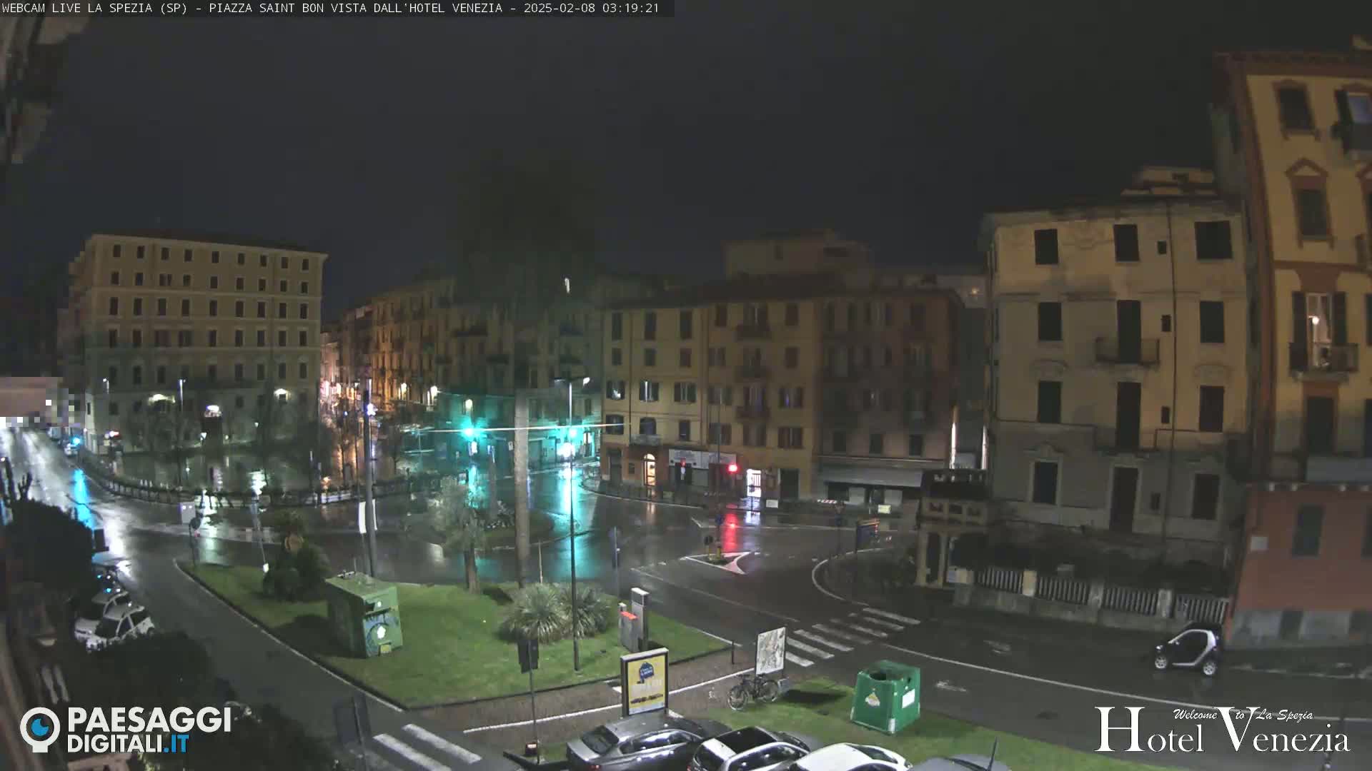 A rain-slicked nighttime city square is surrounded by multi-story buildings, with a few cars visible on the wet streets.