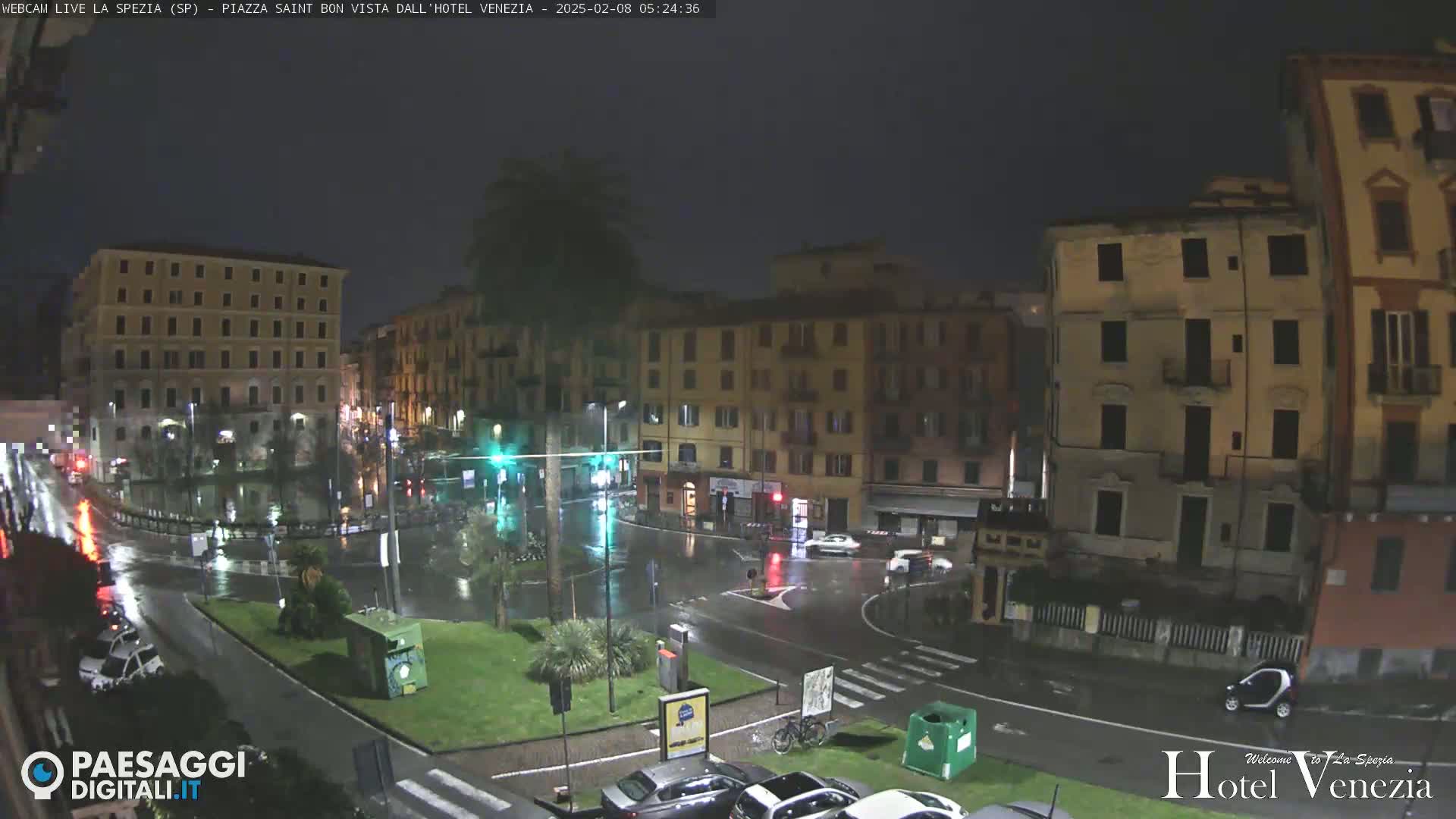 A nighttime view of a rain-soaked European city square, showing several multi-story buildings and sparse traffic under dark skies.