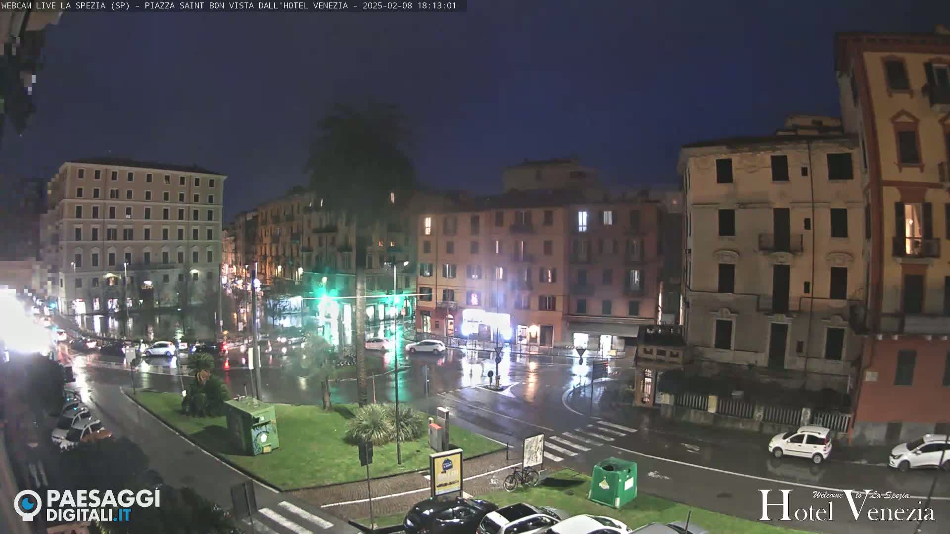 A rain-slicked town square at night, surrounded by multi-story buildings, with cars driving on wet streets.