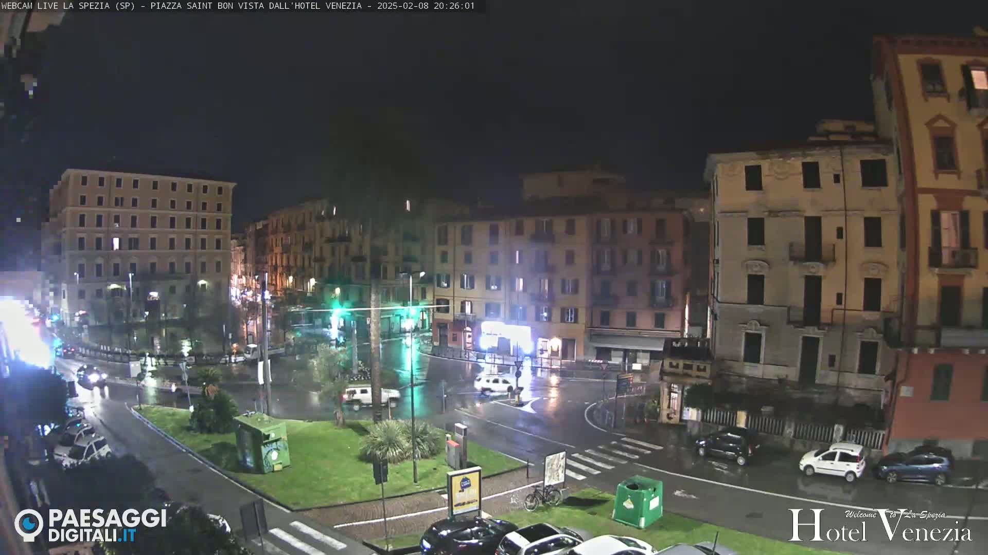 A nighttime view of a rain-slicked city square, surrounded by multi-story buildings, with several cars visible on the wet streets.