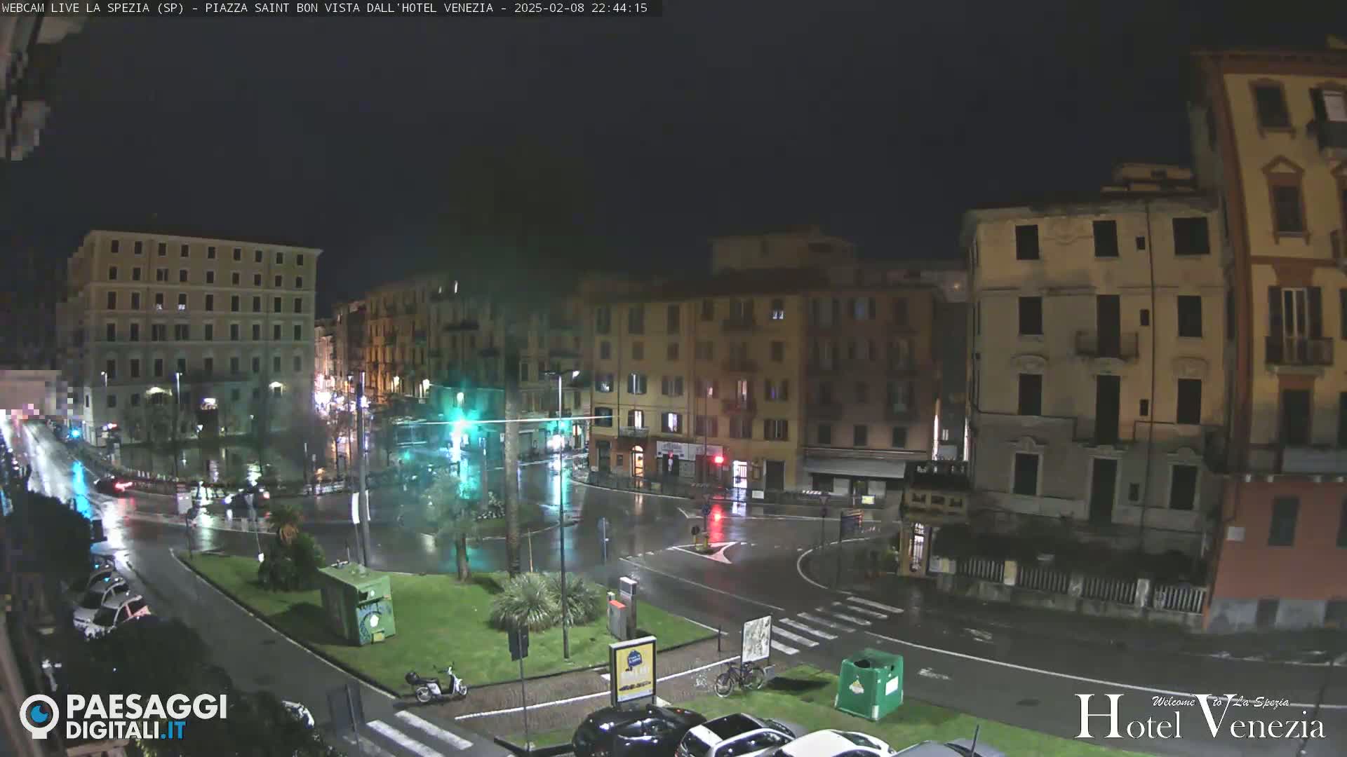 A rain-slicked city square at night, surrounded by multi-story buildings with streetlights illuminating wet roads and parked cars.