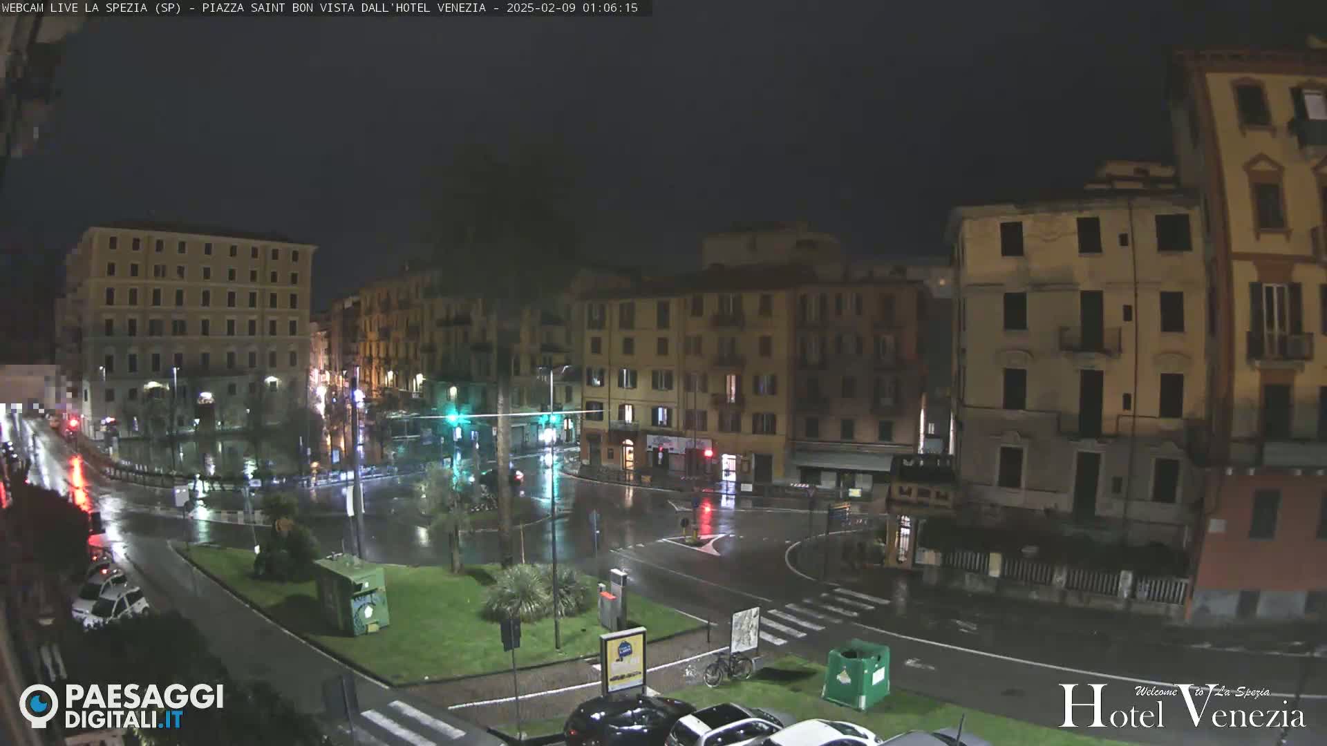 A rain-slicked, nighttime city square is surrounded by multi-story buildings under a dark sky.