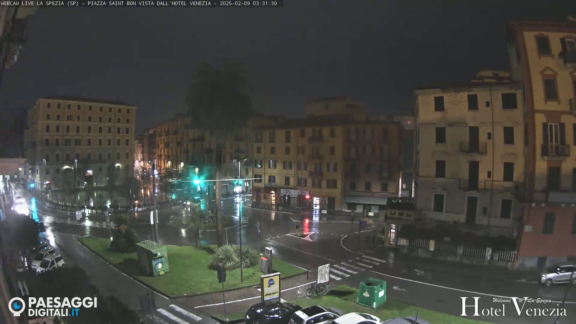 A rain-slicked city square at night, surrounded by multi-story buildings, with a few cars visible on the wet streets.