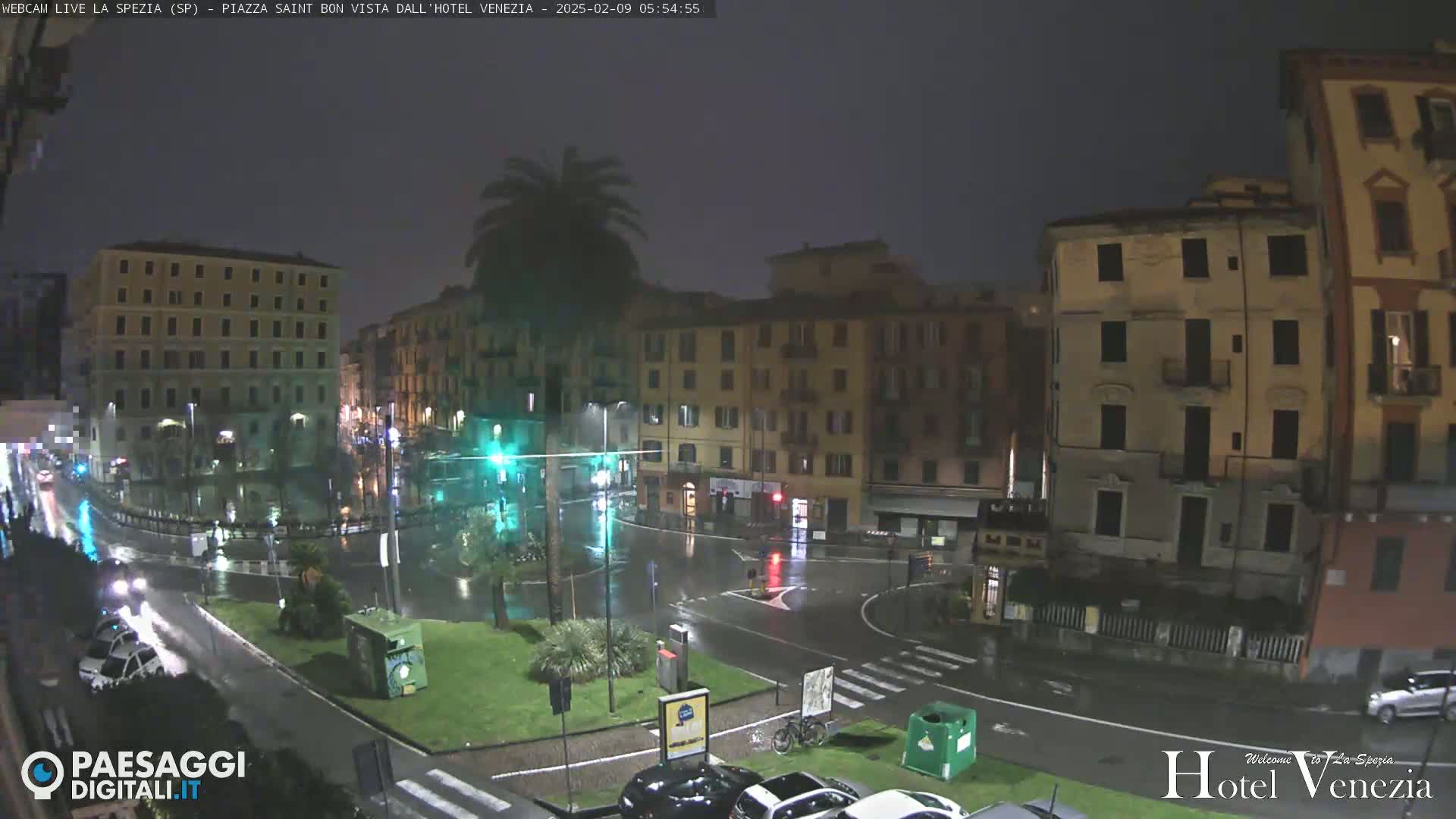 A rain-soaked city square at night, surrounded by multi-story buildings, with a few cars visible on wet roads.