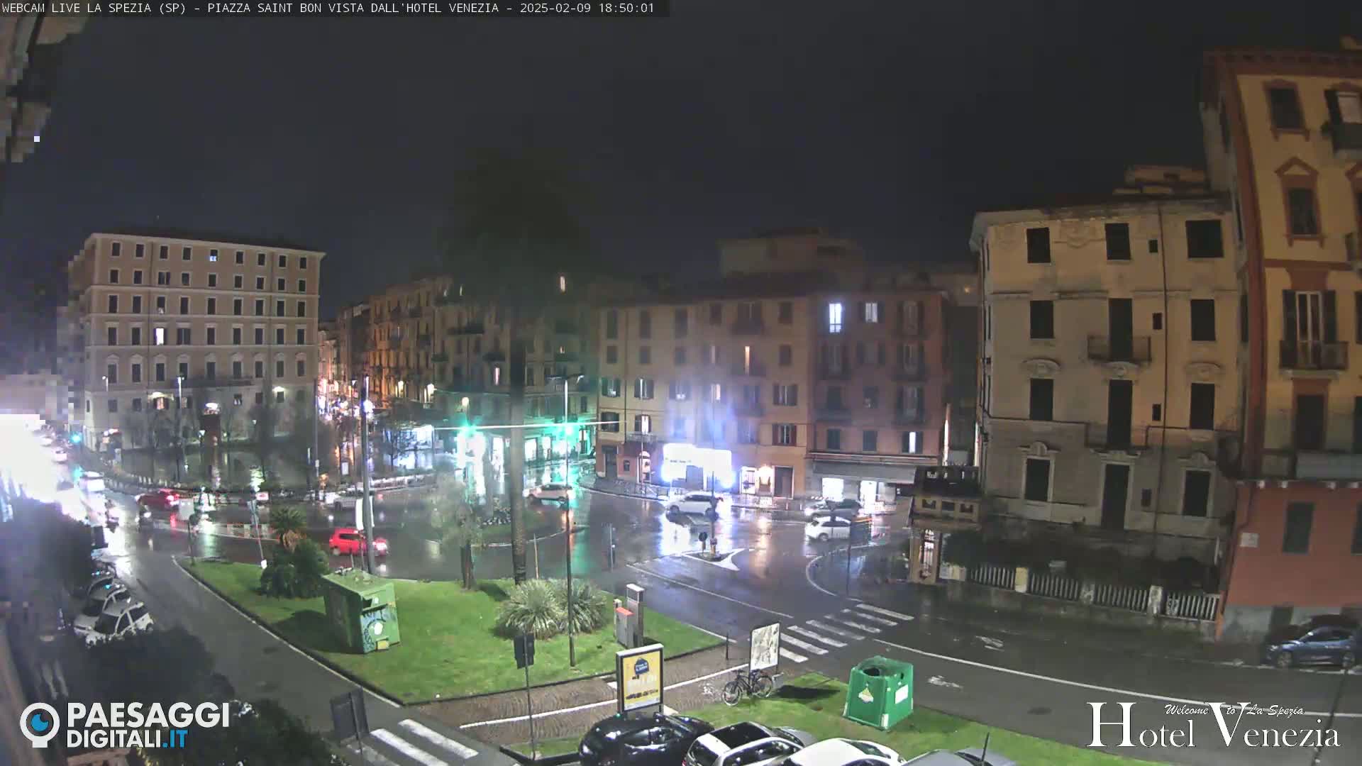 A rain-slicked town square at night, illuminated by streetlights, shows several multi-story buildings and sparse traffic.