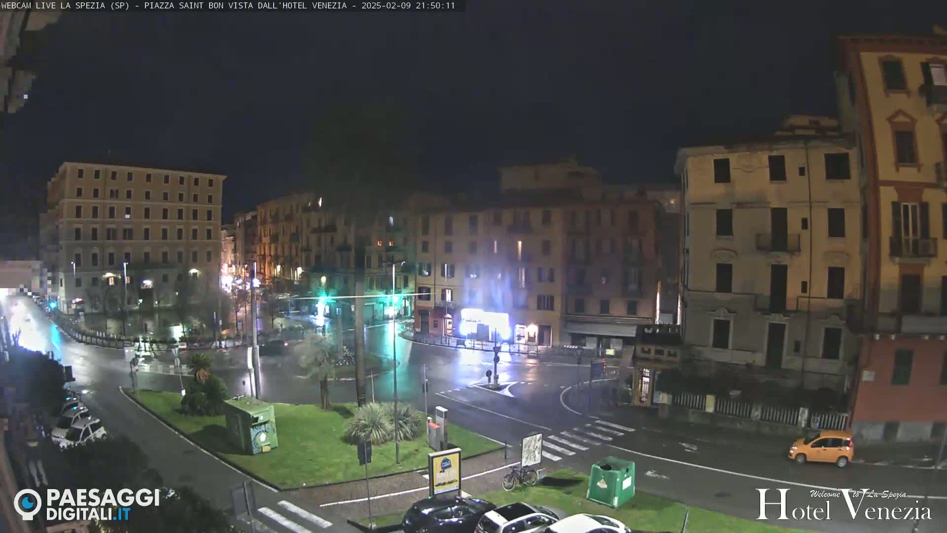 A rain-slicked, nighttime city square is surrounded by multi-story buildings, with a few cars and a bicycle visible.