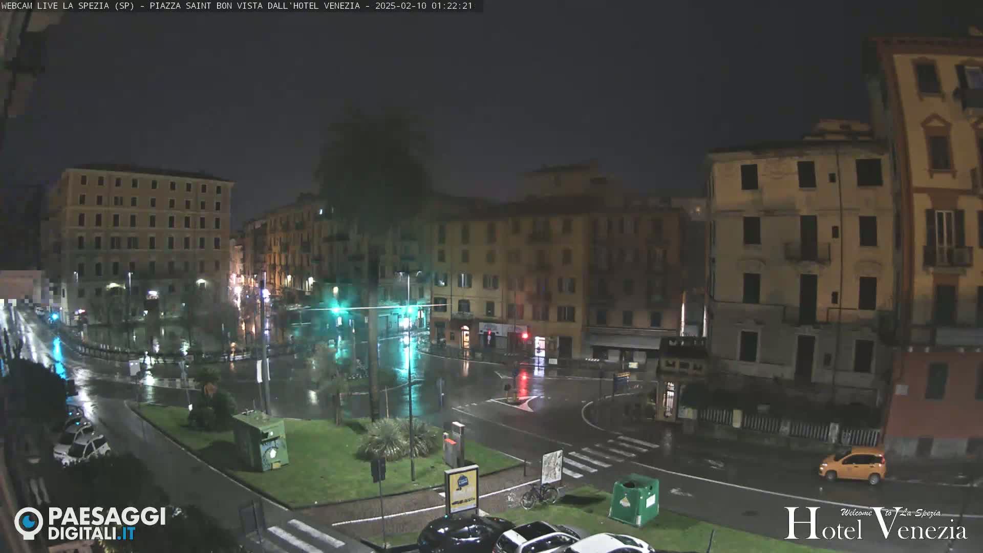 A rain-slicked, nighttime city square is surrounded by multi-story buildings, with a few cars visible in the wet streets.