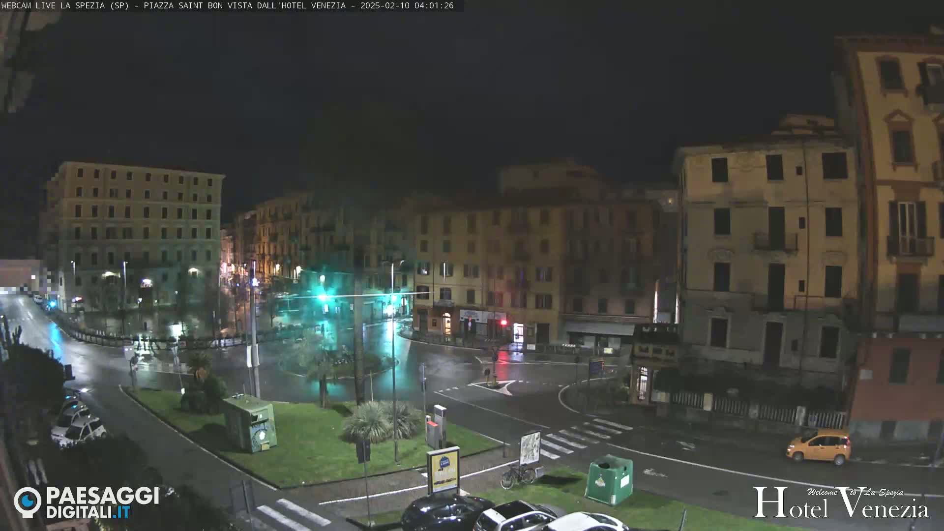 A nighttime, rainy view of a city square with several buildings surrounding a roundabout.