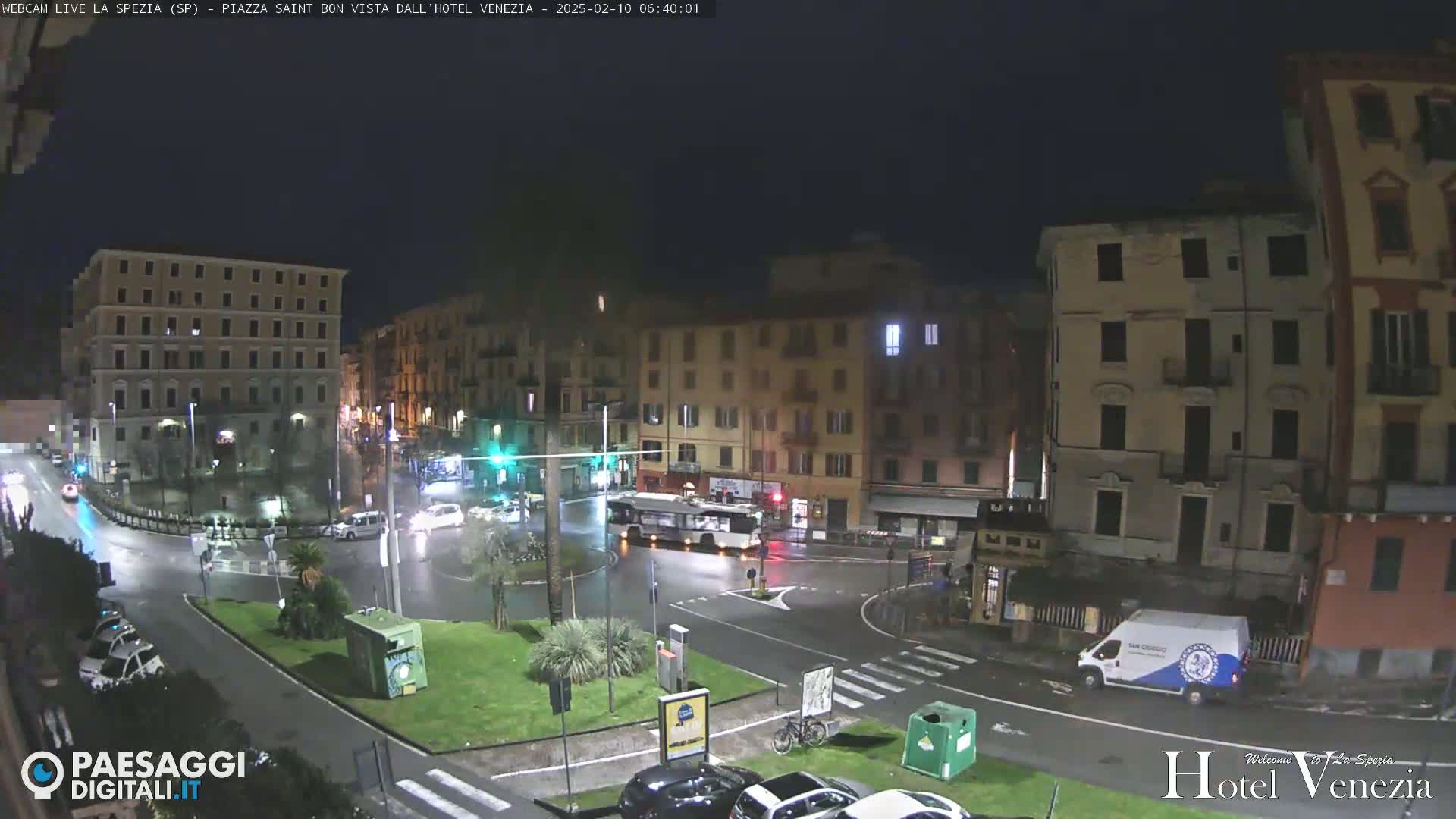 A nighttime view of a wet, city square with several buildings, cars, and a bus visible under dark, overcast skies.