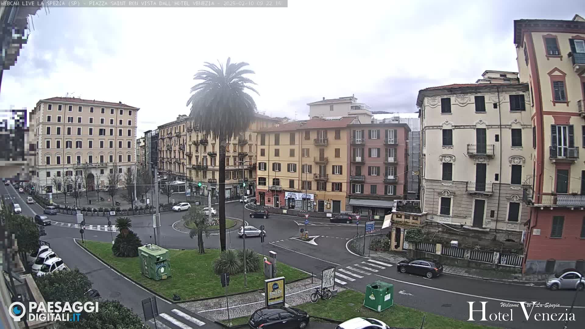 A mostly cloudy day in a European city square with several multi-story buildings surrounding a roundabout with cars and a tall palm tree in the center.