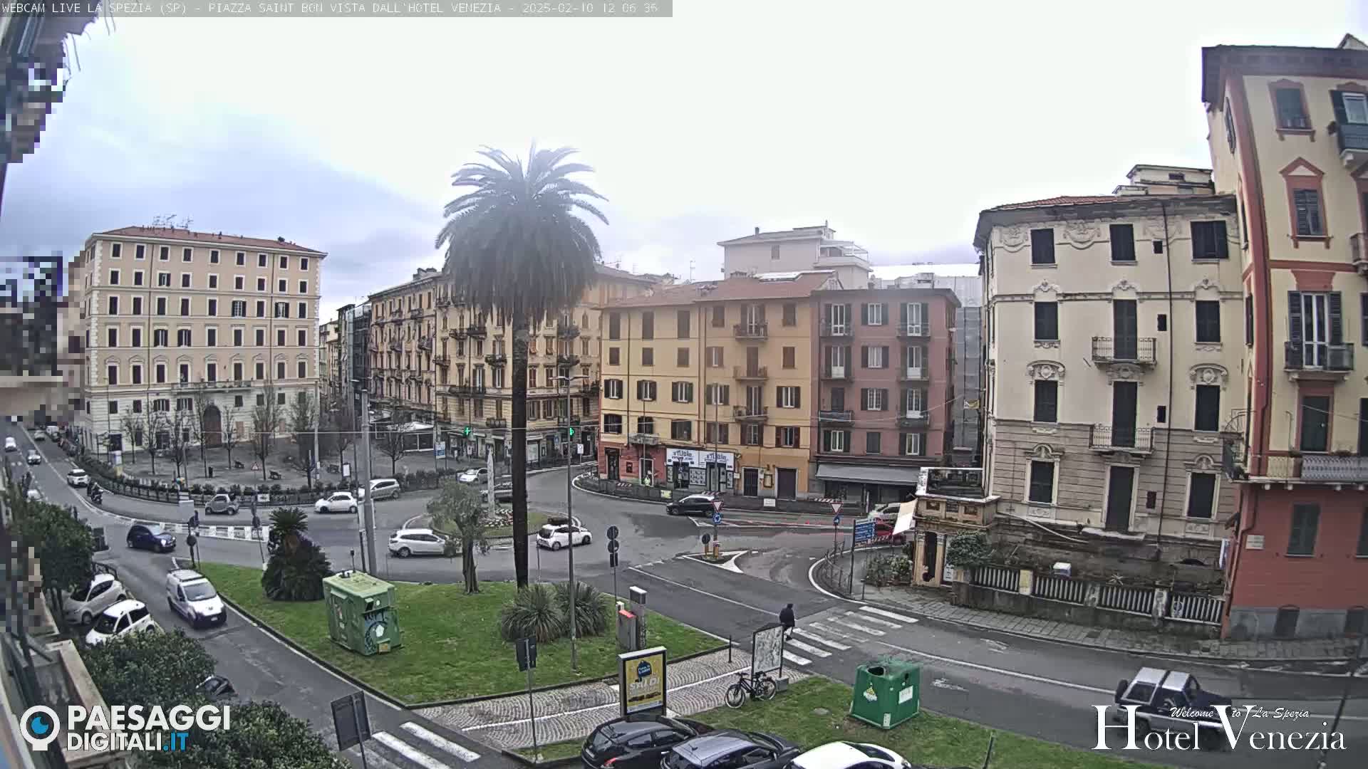 A mostly cloudy day in a city square with several multi-story buildings surrounding a roundabout, where cars are driving, and a palm tree is centered in the square.