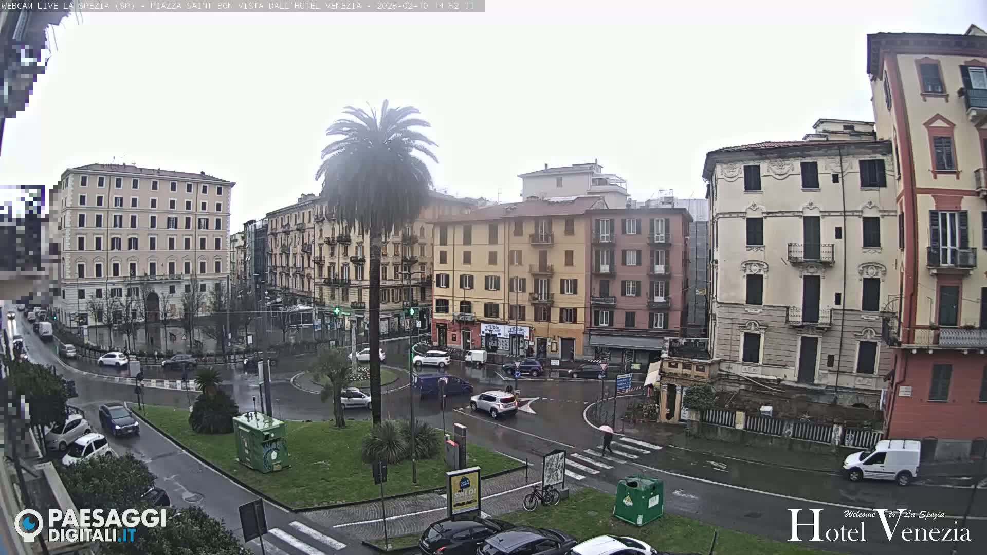 A rainy day scene shows a city square with several multi-story buildings surrounding a roundabout where cars are driving.