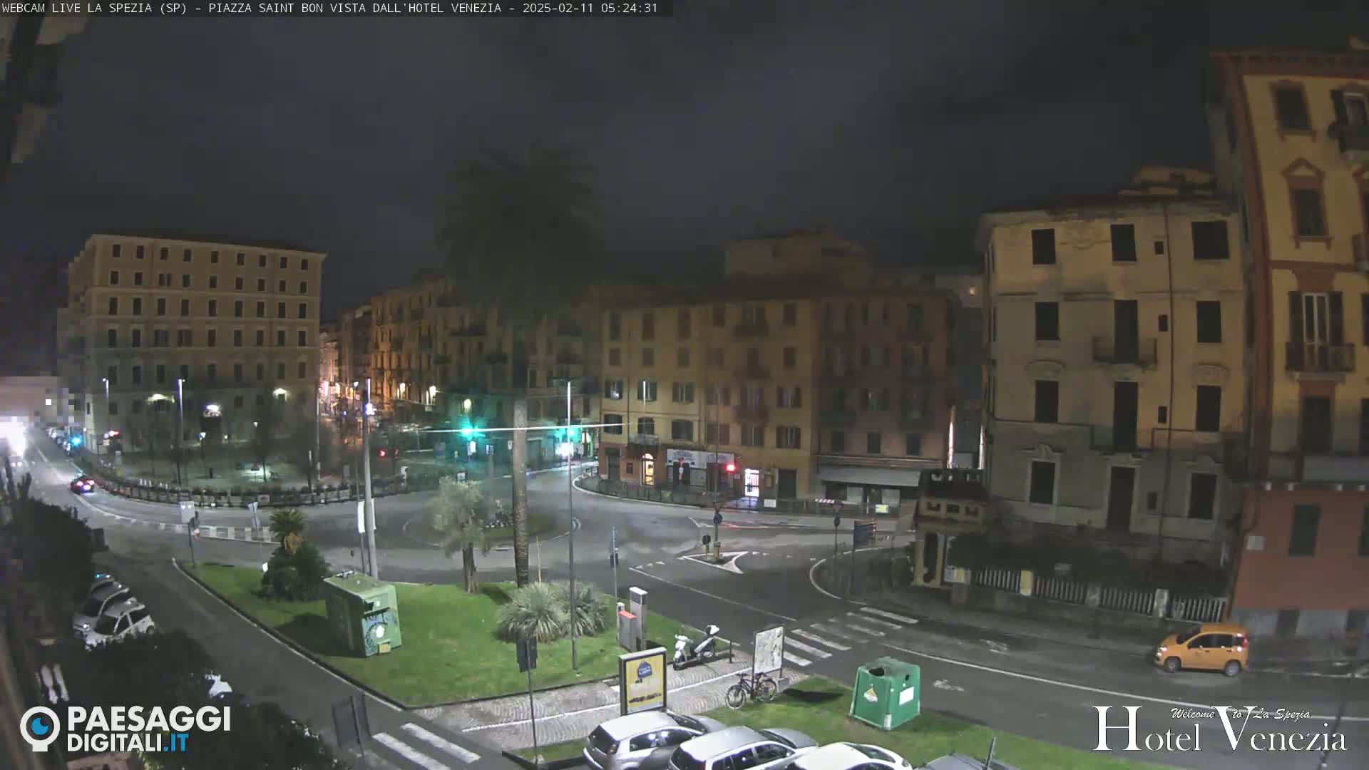 A nighttime view of a town square, with several multi-story buildings surrounding a circular roadway, and a few cars parked and driving.