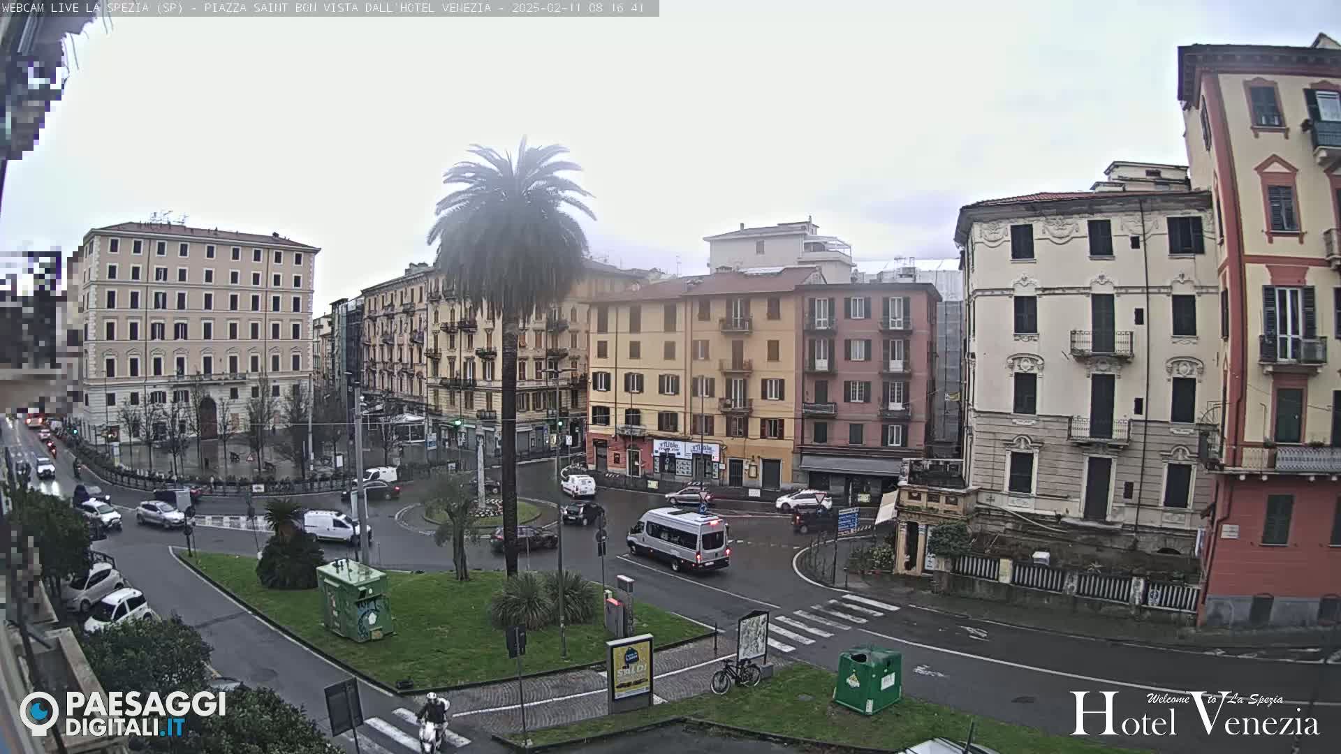 A mostly cloudy day reveals a city square with several multi-story buildings, a traffic circle with cars and a van, a palm tree, and a bicycle.