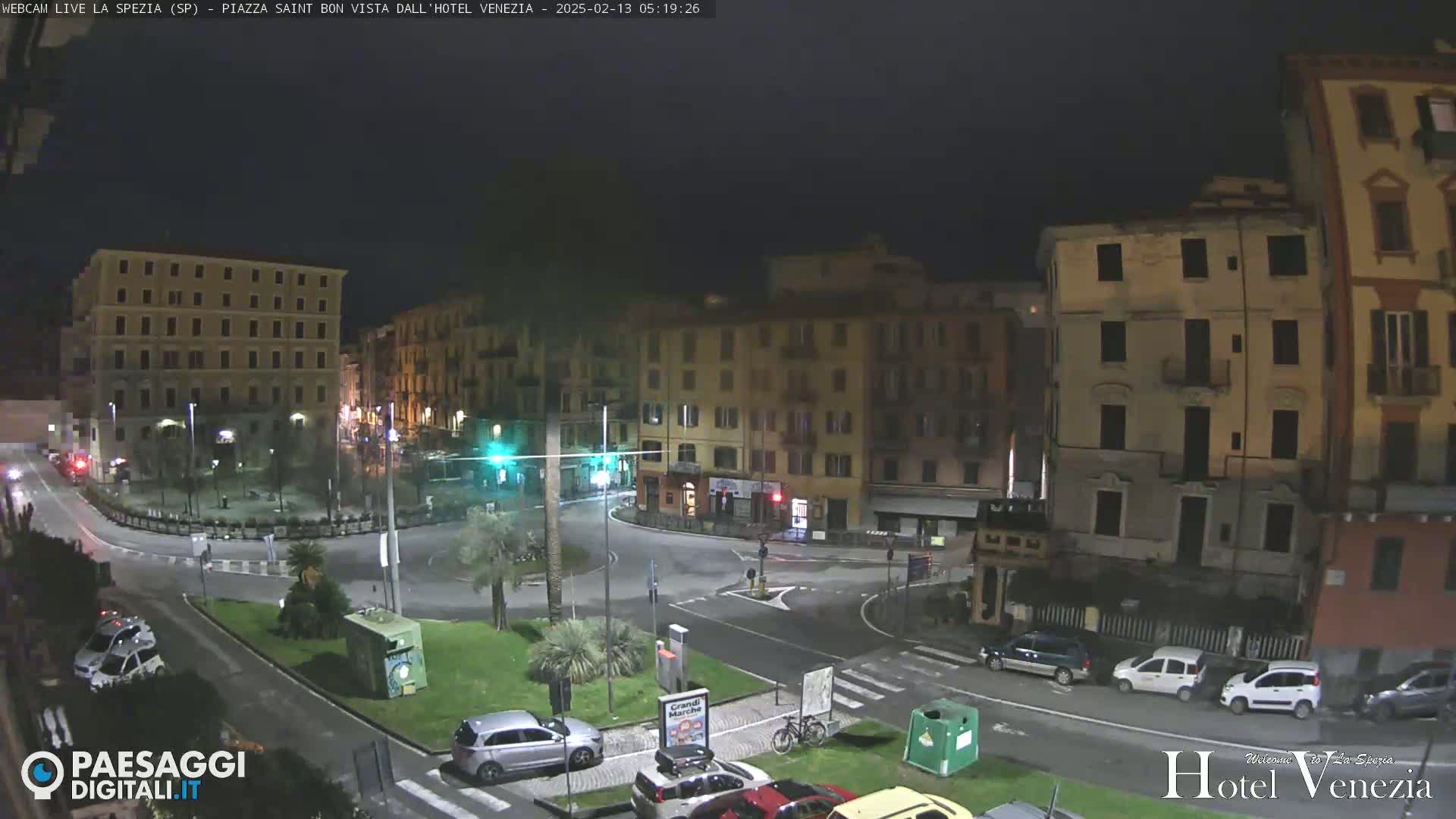 A nighttime view of a city square with several buildings, cars parked along the streets, and a small, planted area in the center of the square.