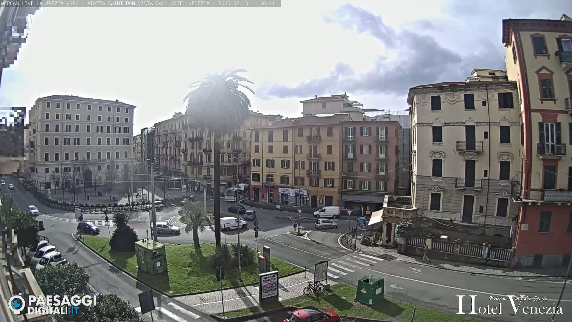 A mostly sunny day reveals a city square with several multi-story buildings, a roundabout, and sparse traffic.