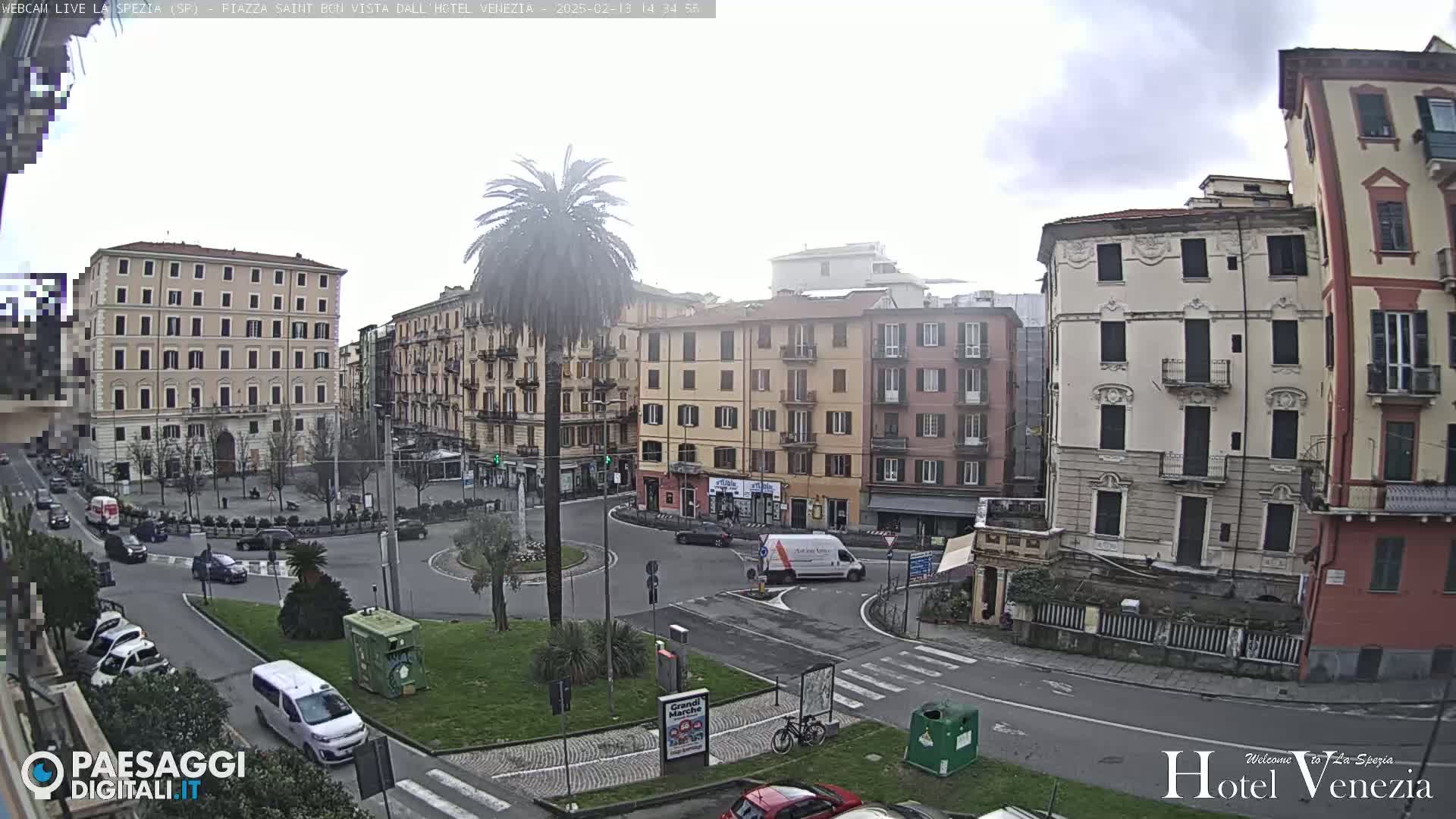 A mostly cloudy day in a European city square shows several multi-story buildings surrounding a traffic circle with cars and a lone palm tree in the center.