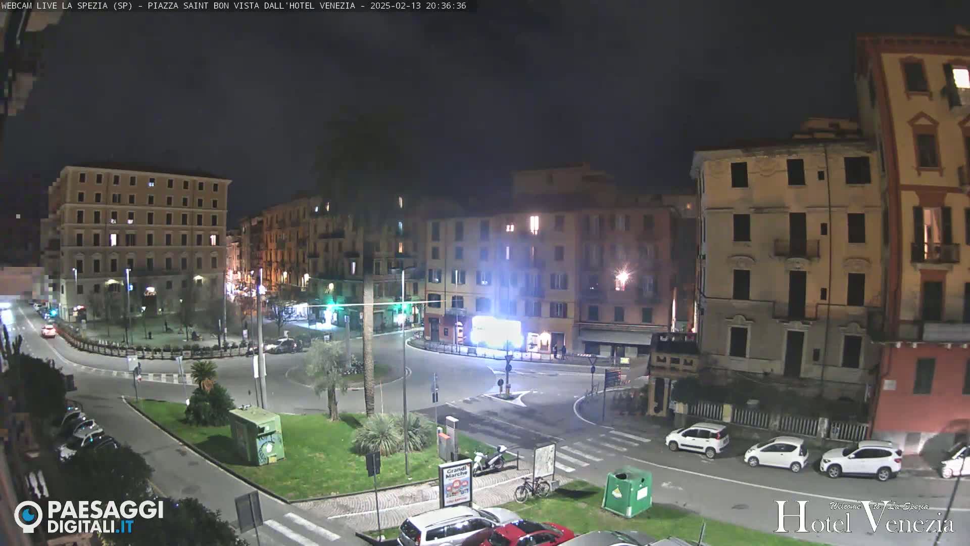 A nighttime view of a mostly empty town square surrounded by multi-story buildings, with several cars parked along the edges and a few cars driving in the square.