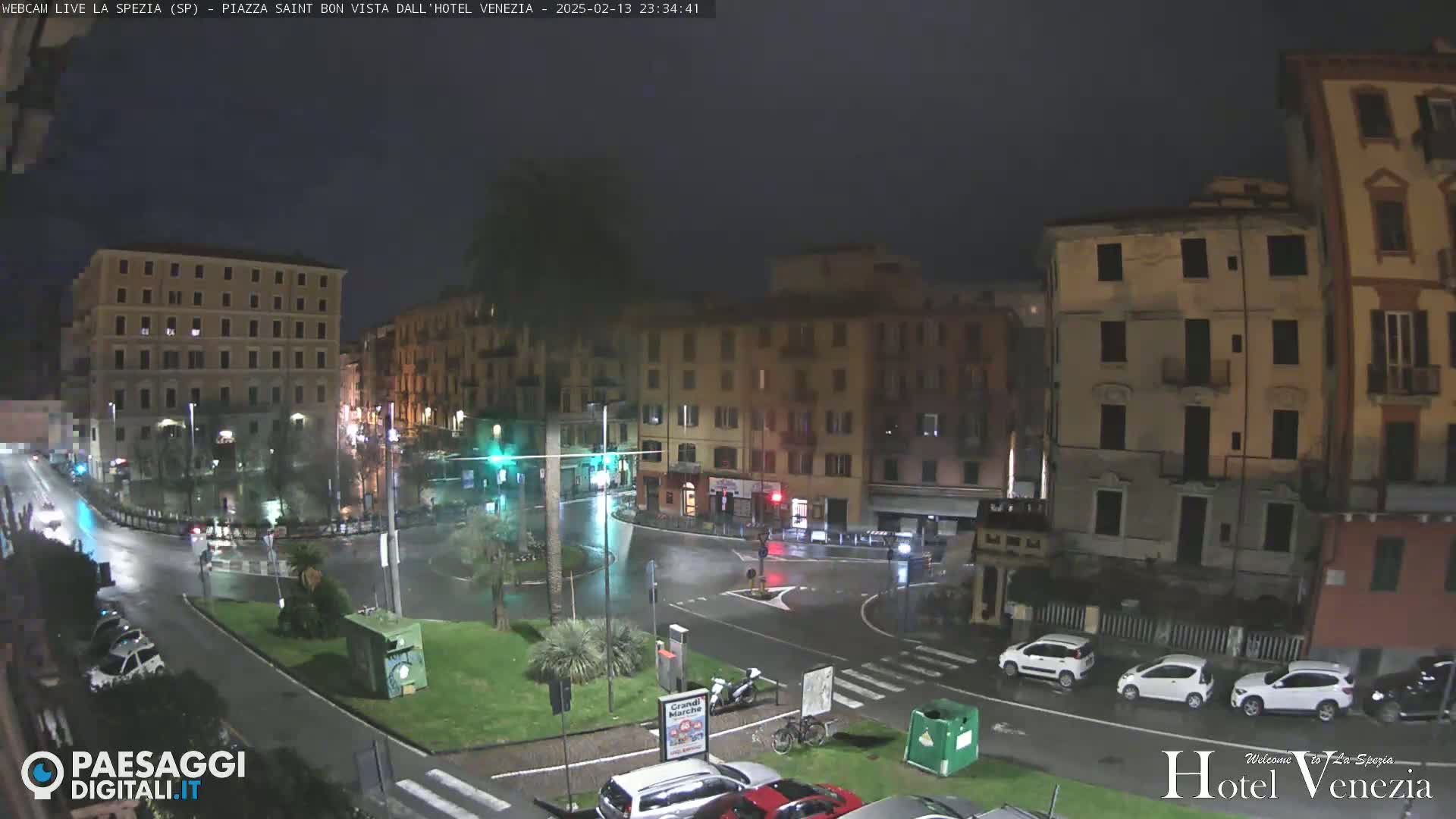 A rain-slicked, nighttime city square is surrounded by multi-story buildings, with several cars parked and driving through.
