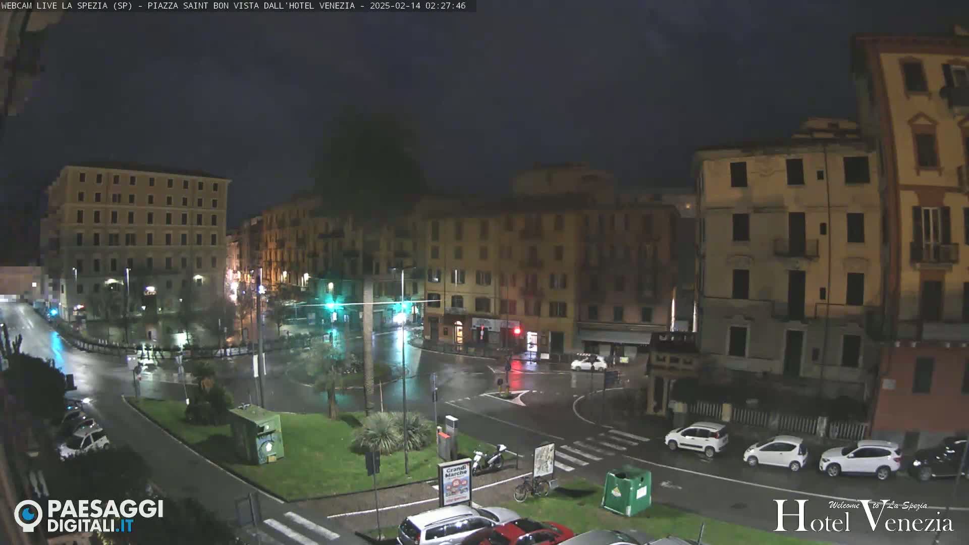A rain-slicked nighttime city square in Italy, surrounded by multi-story buildings, with several cars parked and a few driving through the intersection.