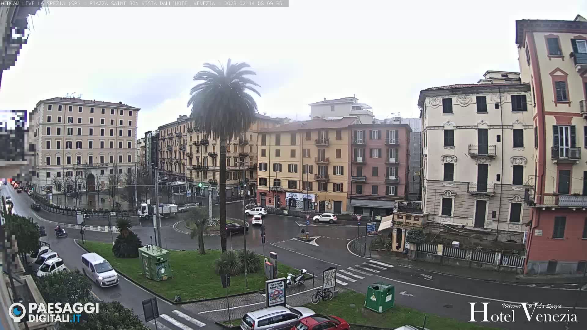 A mostly cloudy day reveals a city square with several multi-story buildings, a palm tree in a roundabout, and light traffic.
