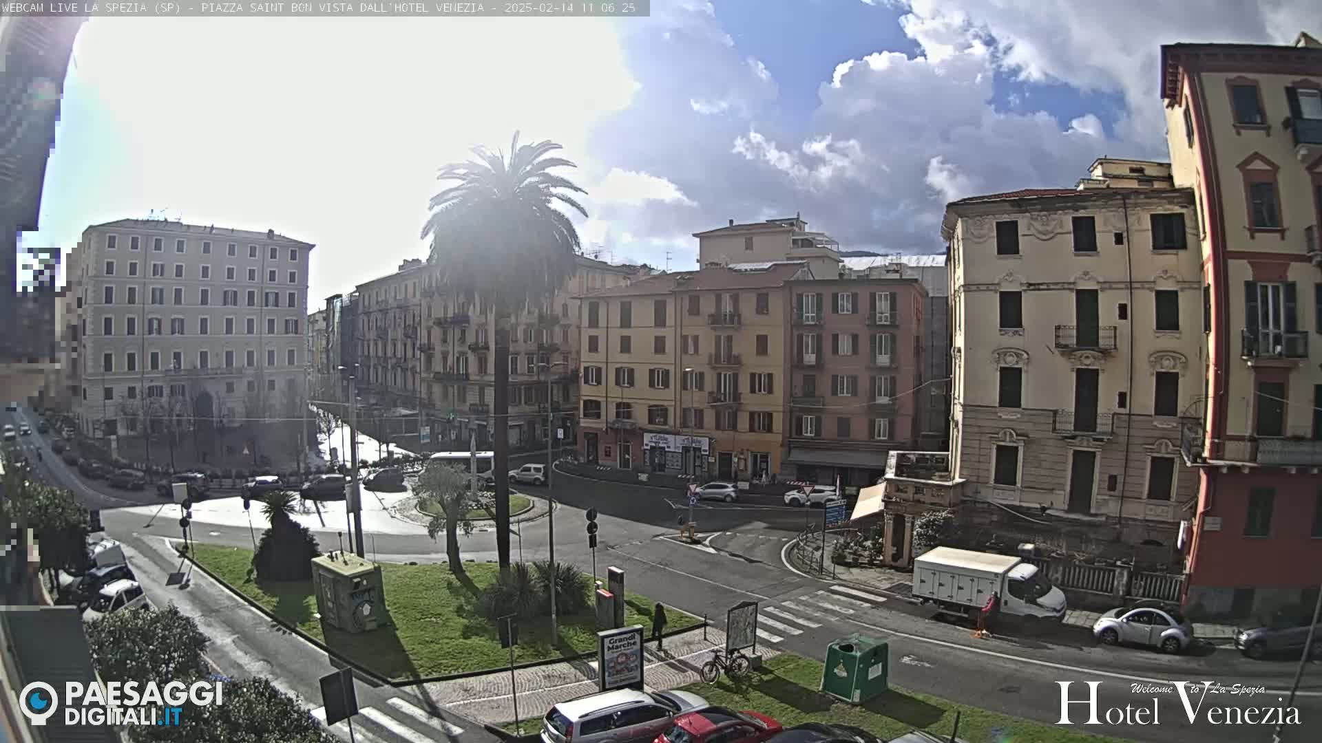 A sunny day reveals a European-style town square with several multi-story buildings, a roundabout, cars, and a palm tree in the center.