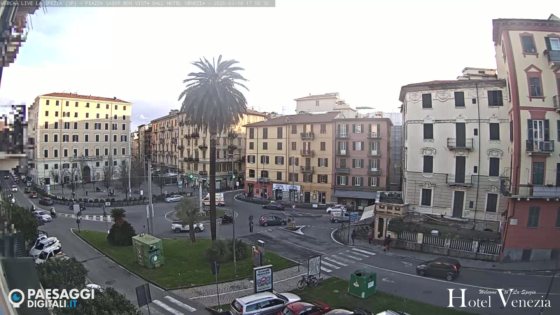 A mostly sunny city square with several multi-story buildings, a palm tree, and sparse traffic.