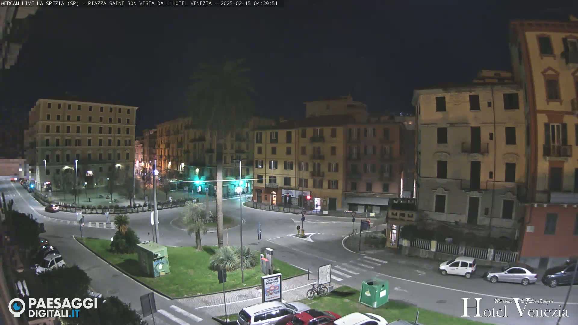 A nighttime, aerial view of a plaza in a city, showing several multi-story buildings, cars parked and driving on surrounding streets, and a small, central park area with a palm tree.