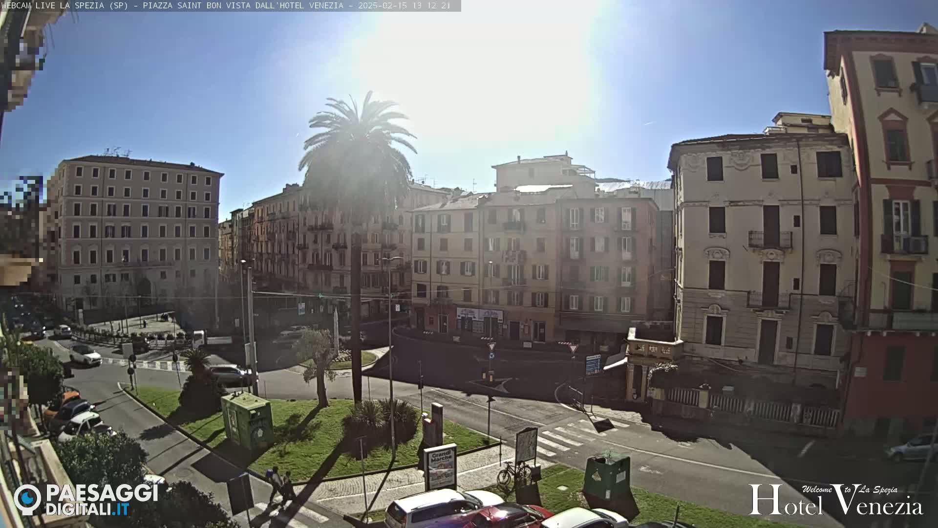 A sunny day in a European city square shows several multi-story buildings surrounding a roundabout with sparse traffic and a few pedestrians.