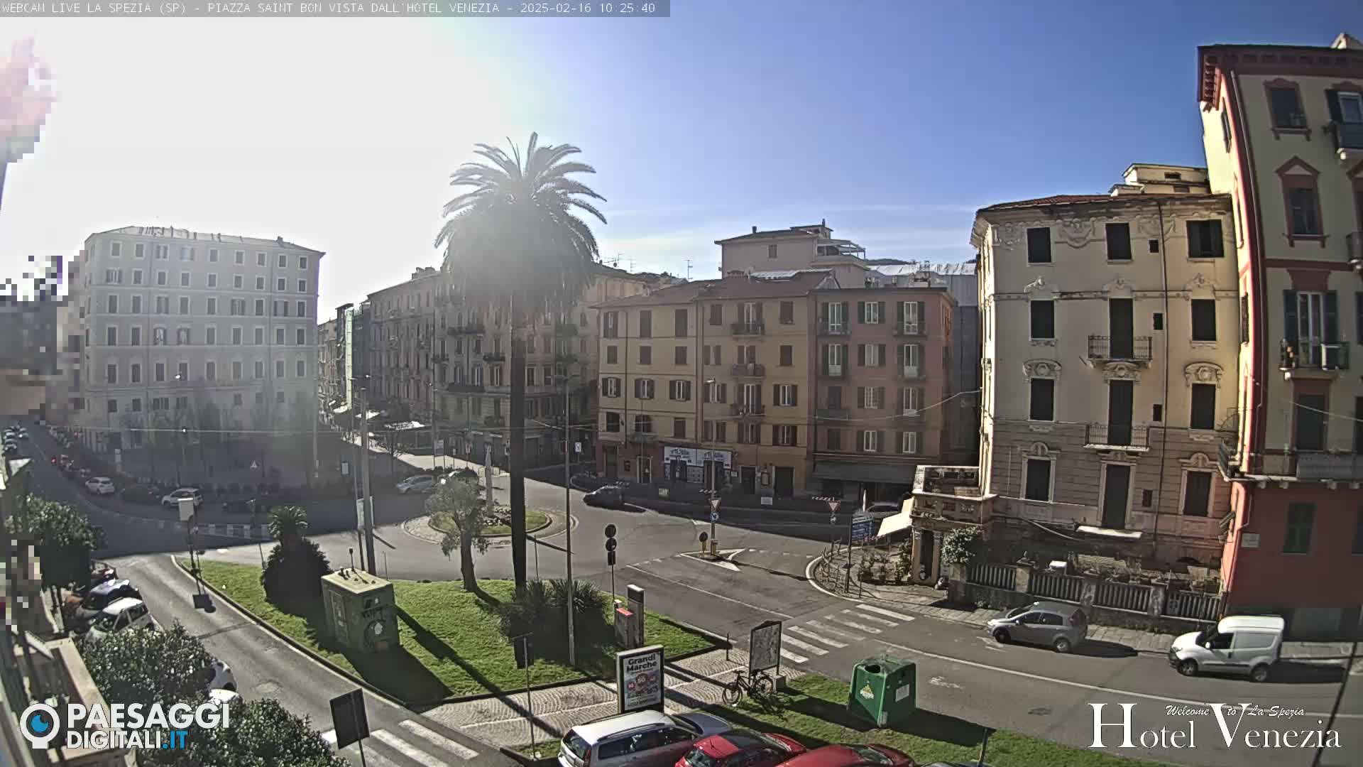A sunny day reveals a city square with several multi-story buildings, palm trees, and sparse traffic.