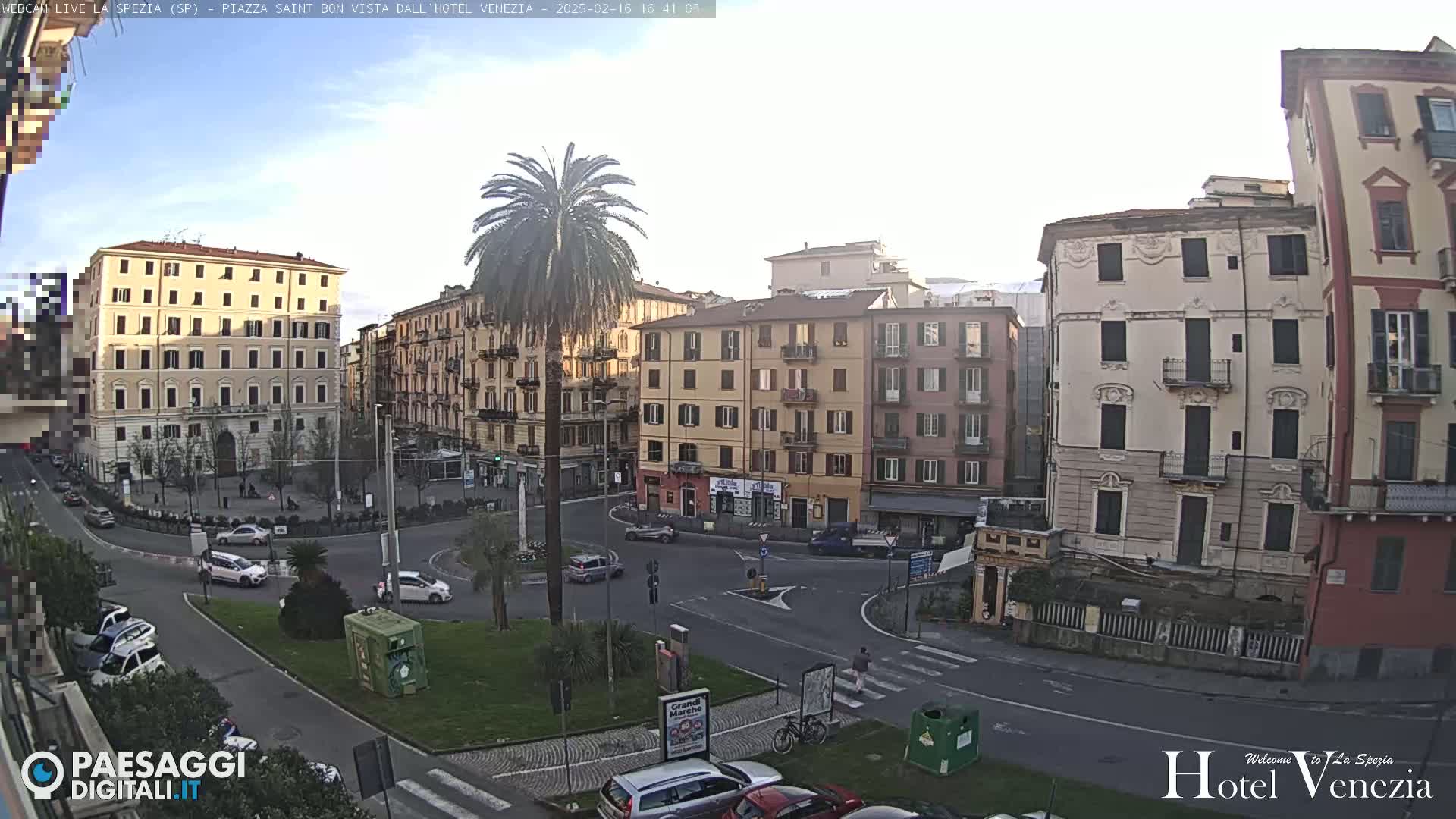 A sunny day reveals a European city square with several multi-story buildings, a palm tree in a circular traffic island, and cars parked along the streets.