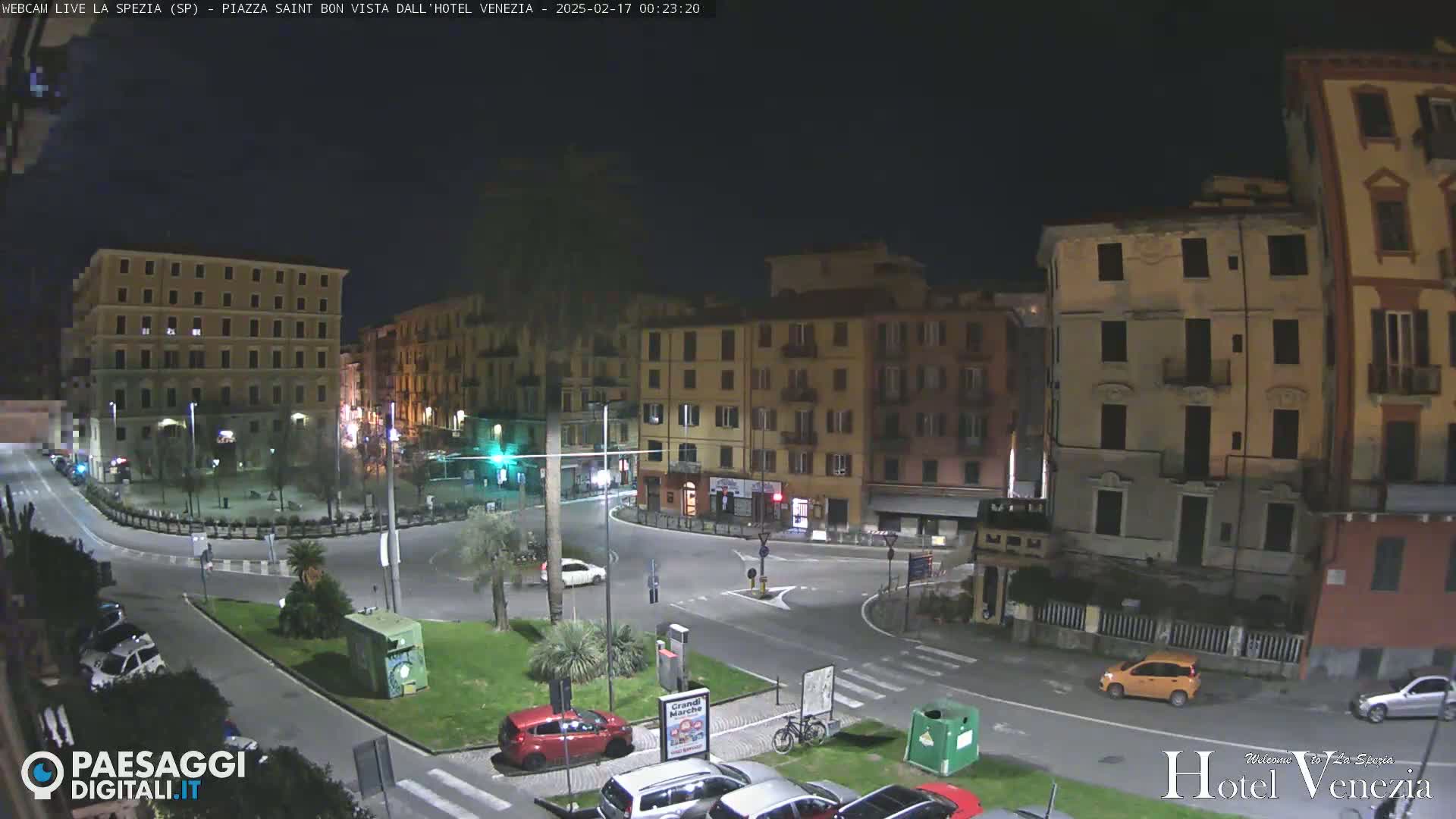 A nighttime view of a city square with several buildings surrounding a roundabout, where cars are parked and driving.