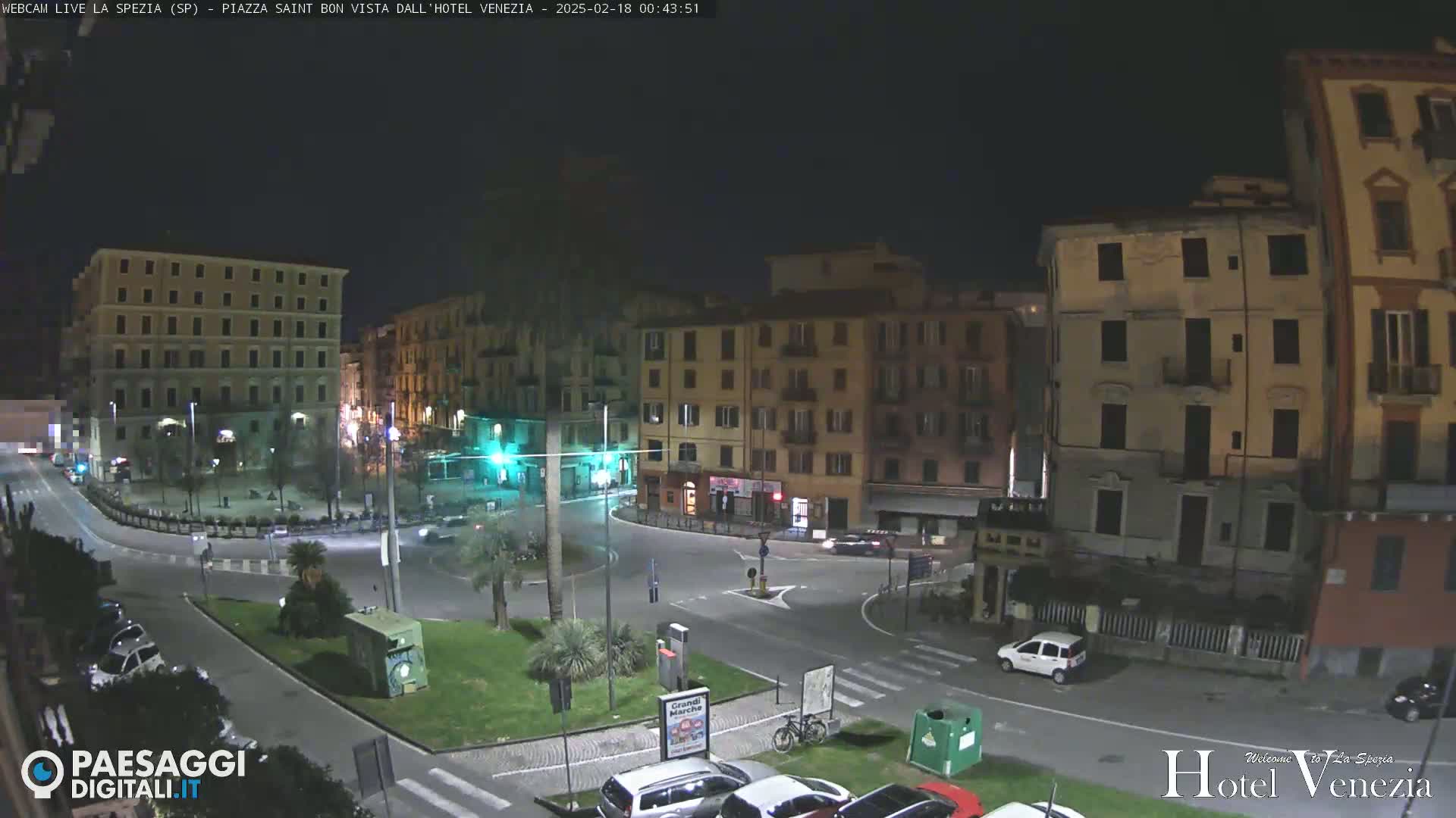 A nighttime, city-center roundabout with several multi-story buildings surrounding it, few cars visible, and a small, sparsely populated plaza at its center.