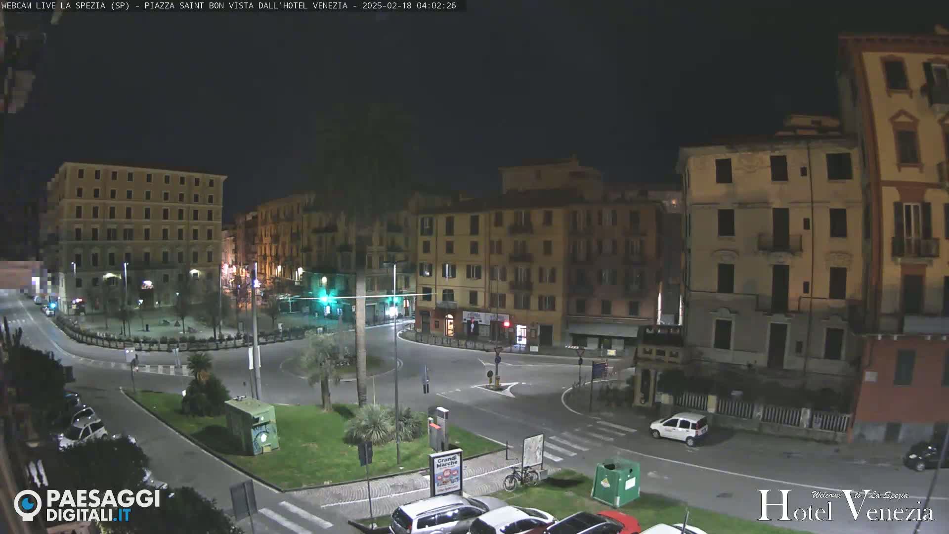 A nighttime view of a city square with several multi-story buildings surrounding a paved area with sparse traffic and small patches of greenery.