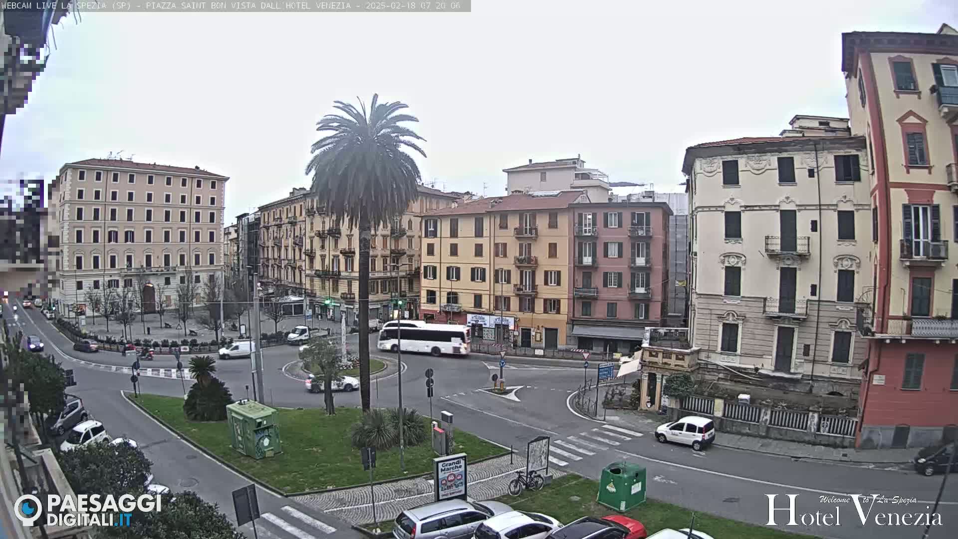 A mostly overcast day reveals a European city square with multi-story buildings surrounding a roundabout where cars and a bus are navigating.