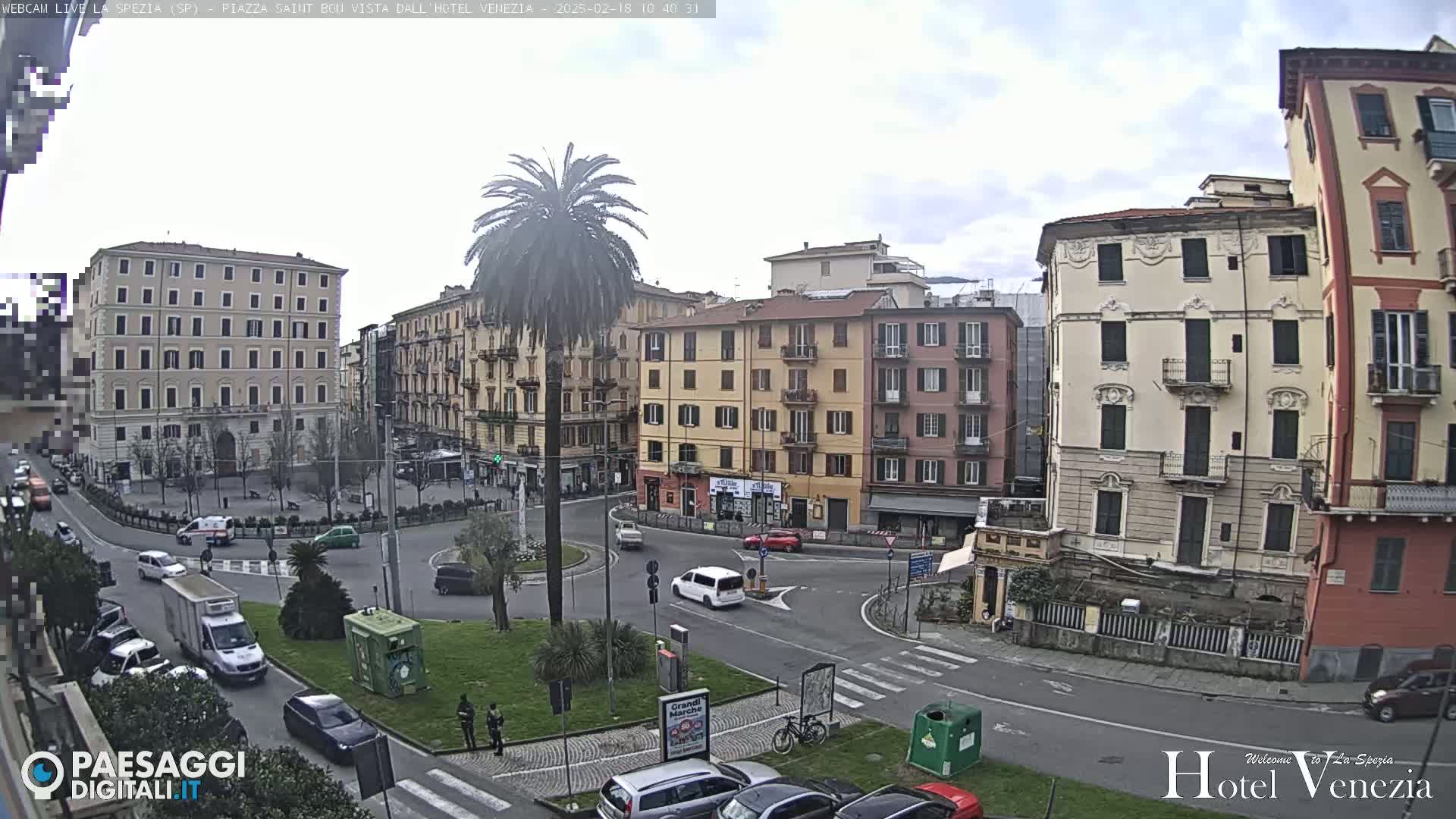 A mostly cloudy day reveals a traffic circle surrounded by multi-story buildings in a European city, centered on a palm tree in a small plaza.