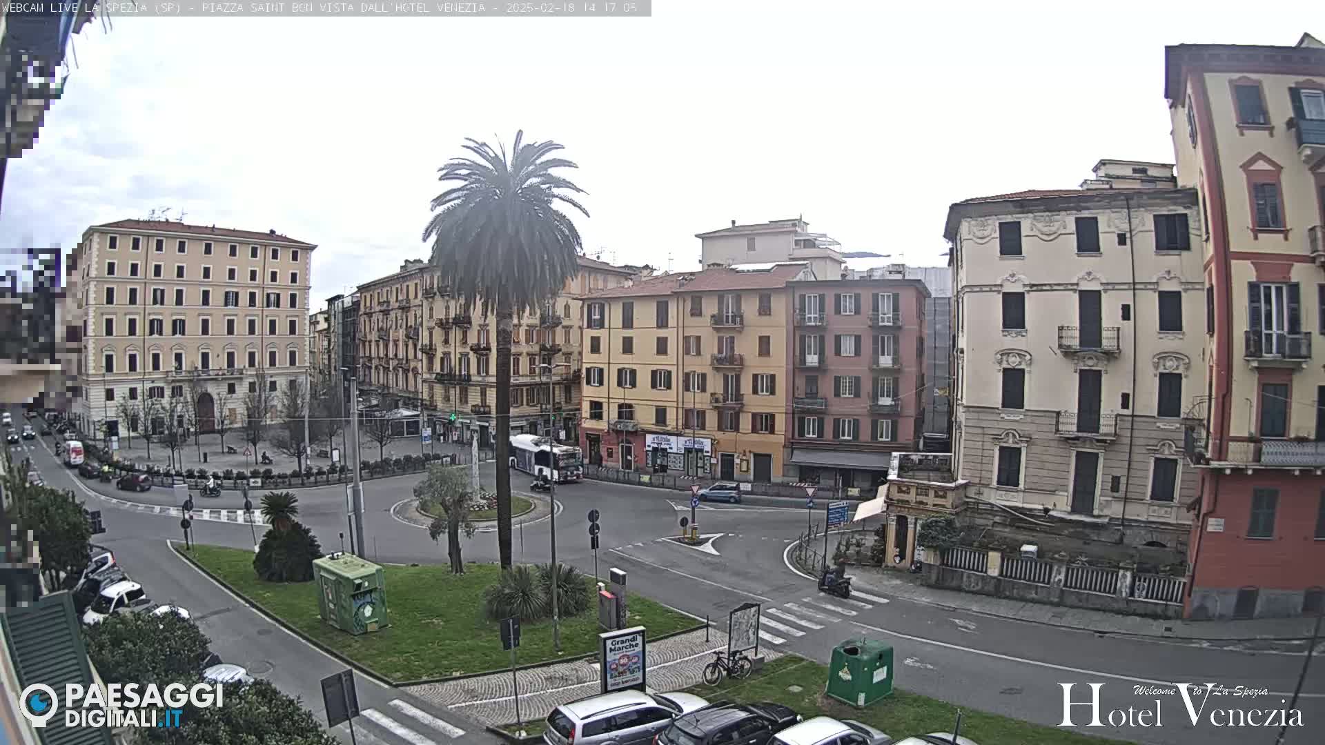 A mostly overcast day reveals a European city square with several multi-story buildings surrounding a traffic circle, a palm tree in the center, and cars parked along the perimeter.