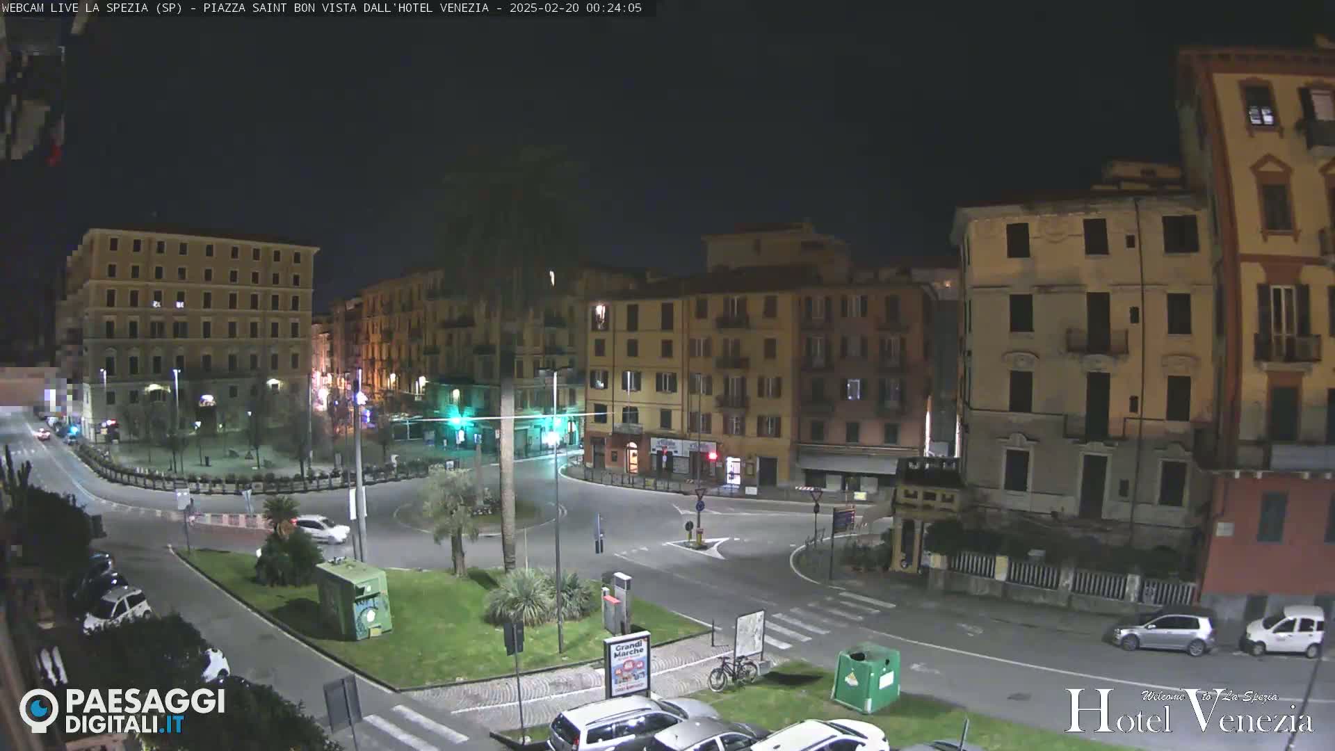 A nighttime view of a city square with several multi-story buildings surrounding a traffic circle, sparse vehicle traffic, and some greenery.