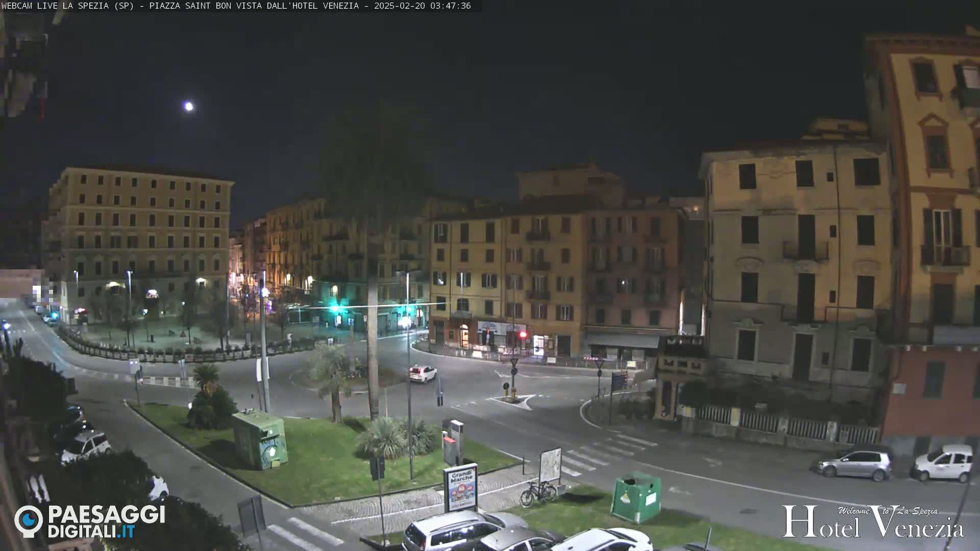 A nighttime view of a mostly empty town square with several multi-story buildings surrounding it under a clear night sky.