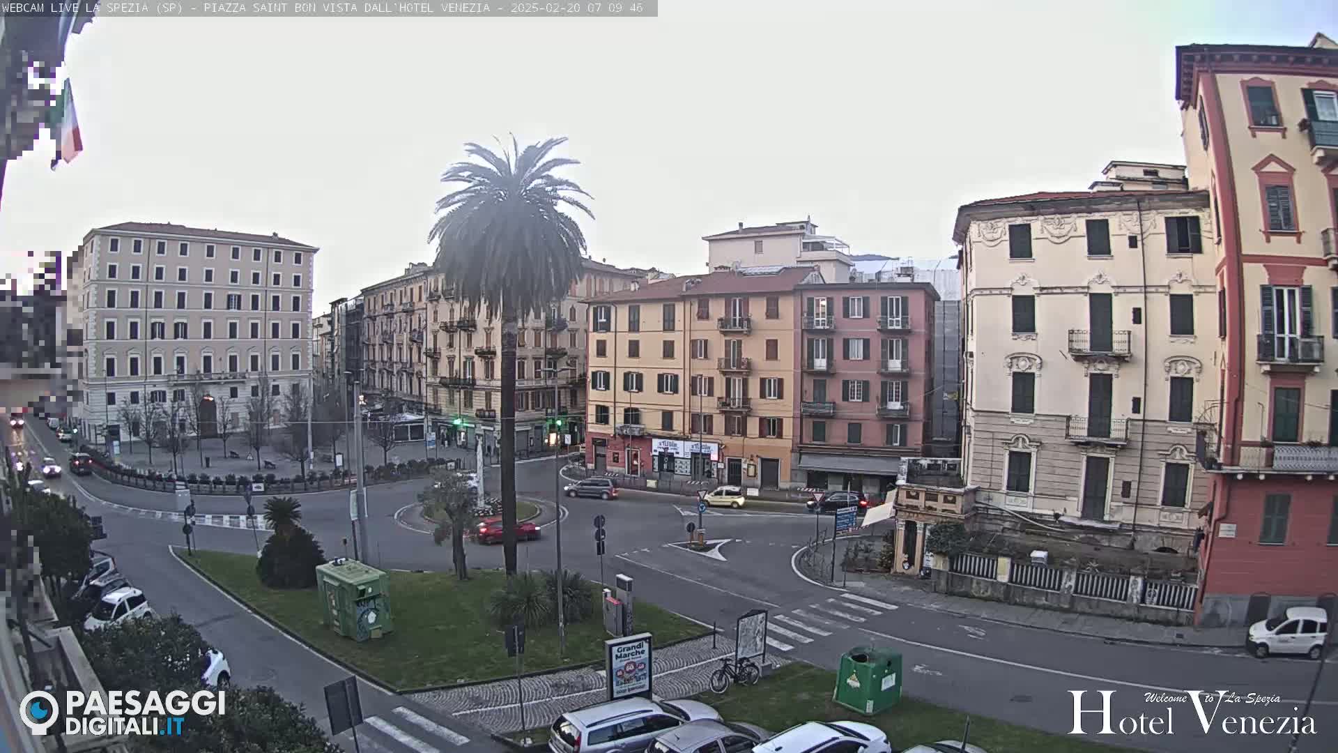 A mostly cloudy day reveals a city square with several multi-story buildings surrounding a roundabout with sparse traffic, and a lone palm tree in the center.