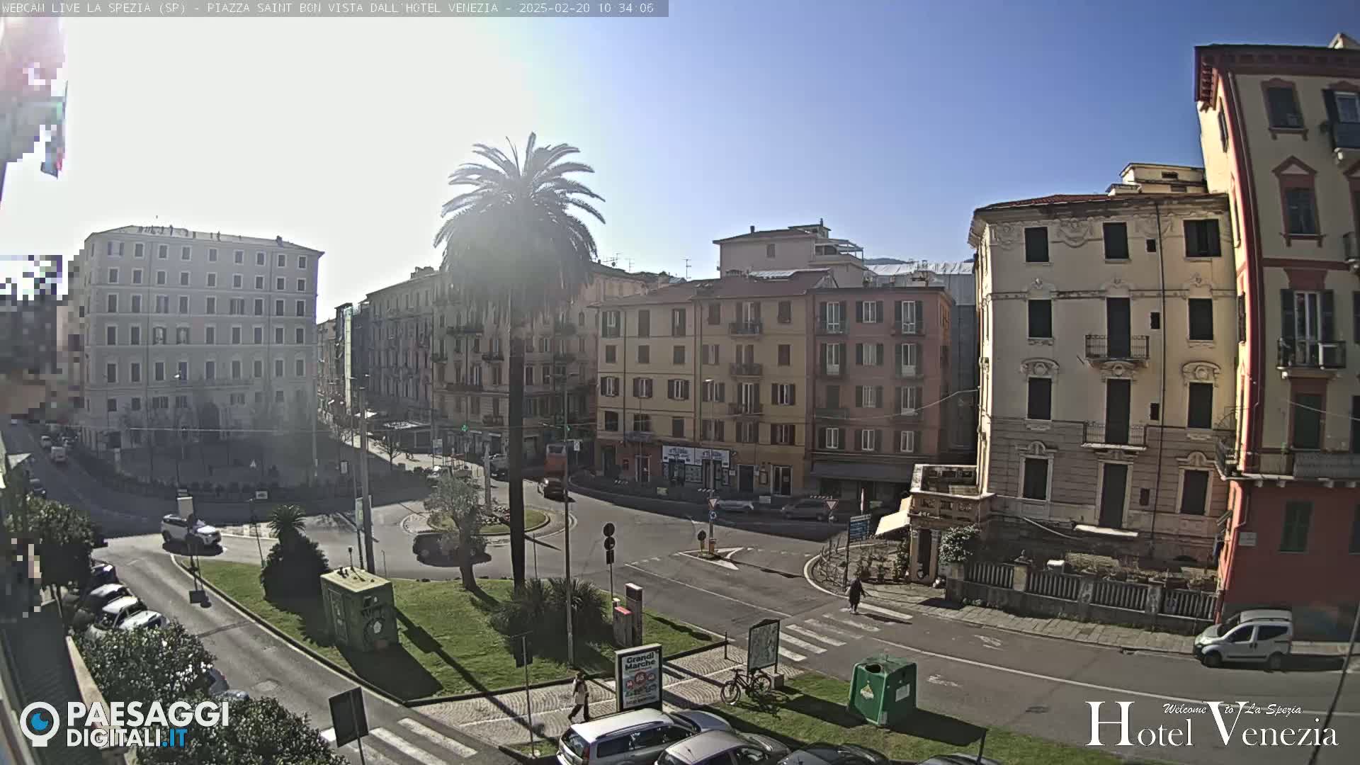 A sunny day in a European city square, showing several multi-story buildings surrounding a roundabout with parked cars and pedestrians.