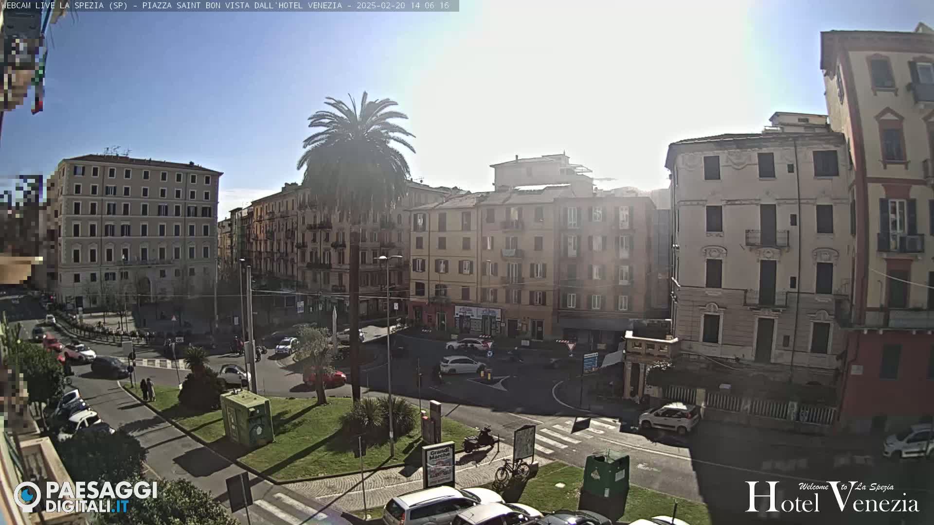 A sunny day in a European city square, showing several multi-story buildings surrounding a traffic circle with cars and pedestrians.