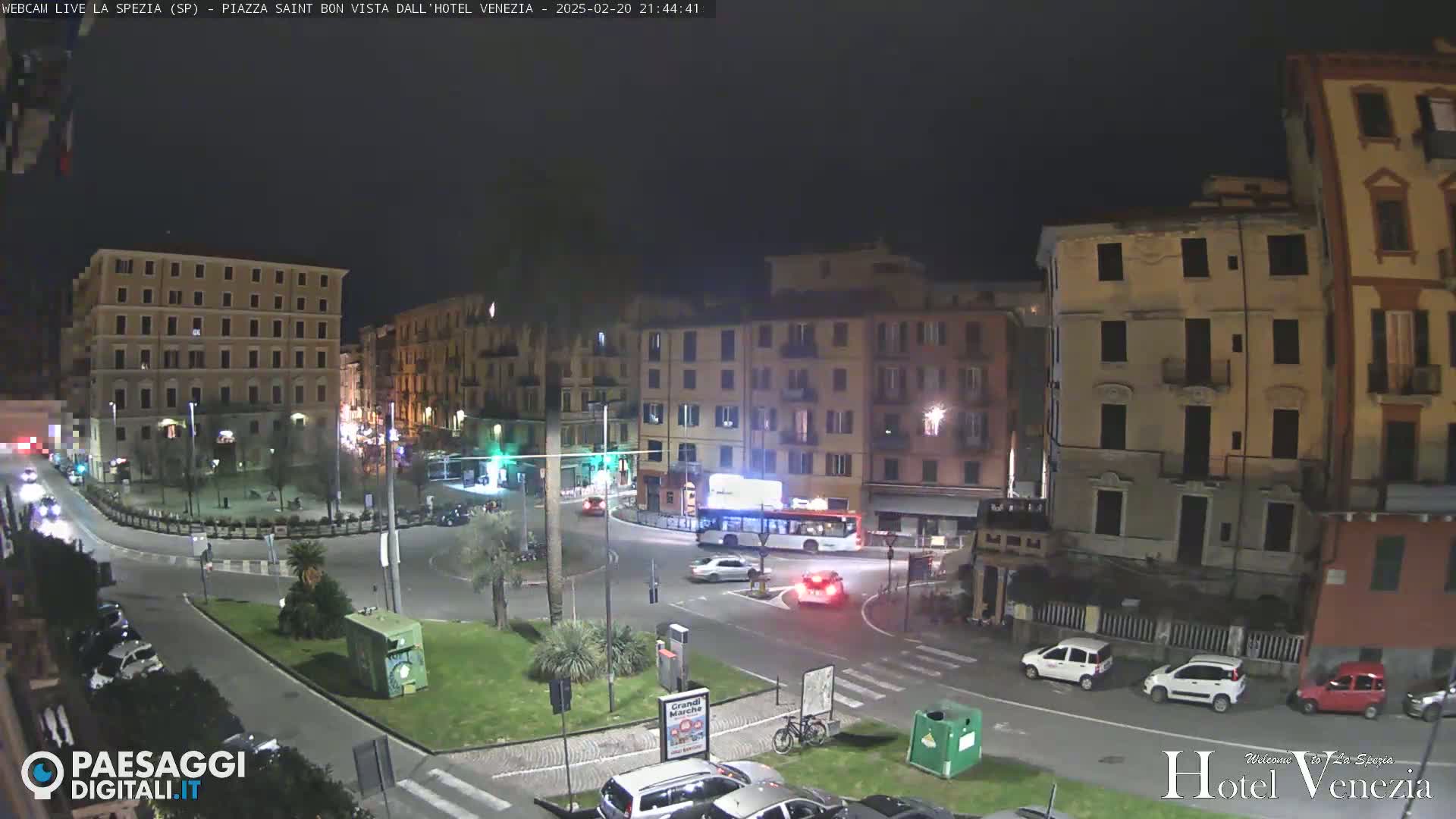 A nighttime view of a city square with several multi-story buildings surrounding it, cars are driving through the square, and there are some palm trees and other plants in the center.