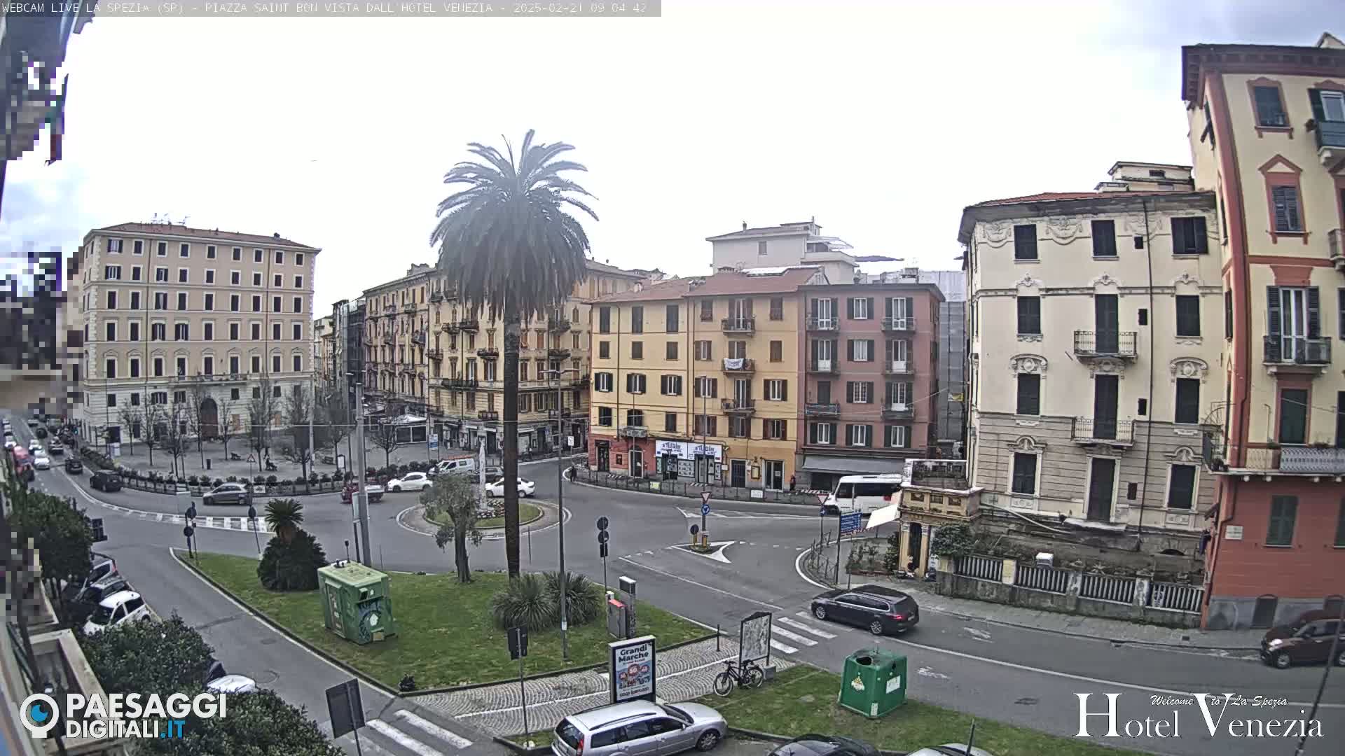 A mostly cloudy day reveals a European city square with several multi-story buildings surrounding a roundabout, where cars are parked and driving, and a tall palm tree stands in the center of a small landscaped area.
