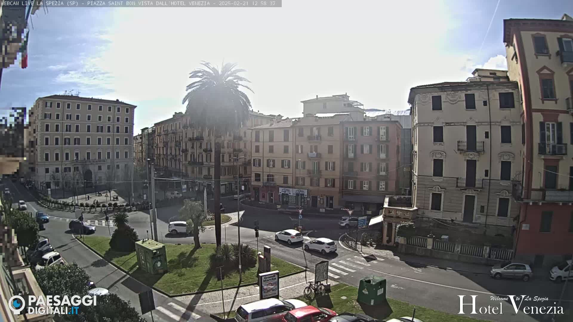 A sunny day in a European city square, showing several multi-story buildings, a roundabout with cars, and a tall palm tree.