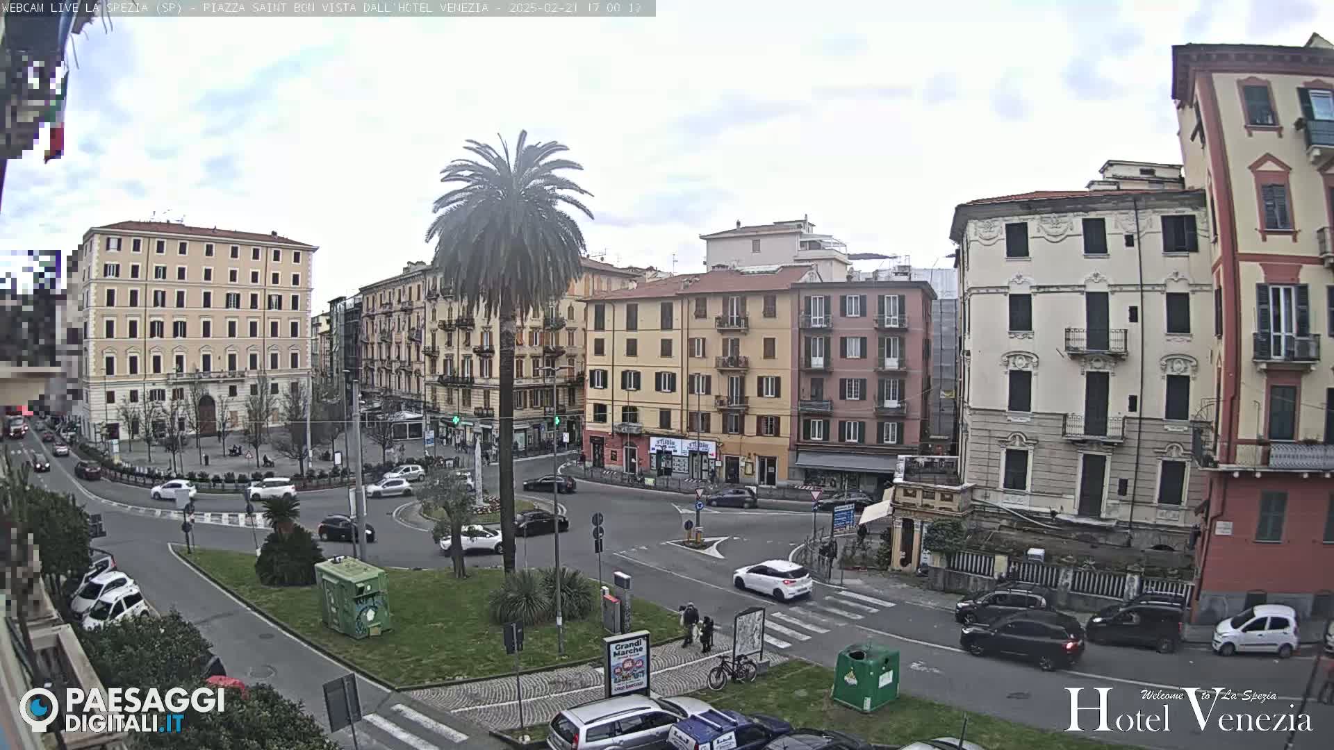 A mostly overcast day shows a traffic circle surrounded by multi-story buildings in a city square, with a palm tree in the center of the circle.